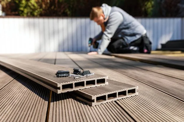A person kneeling on the ground installing brown composite decking boards with screws and a power drill. A gray fence and some bushes are in the background.