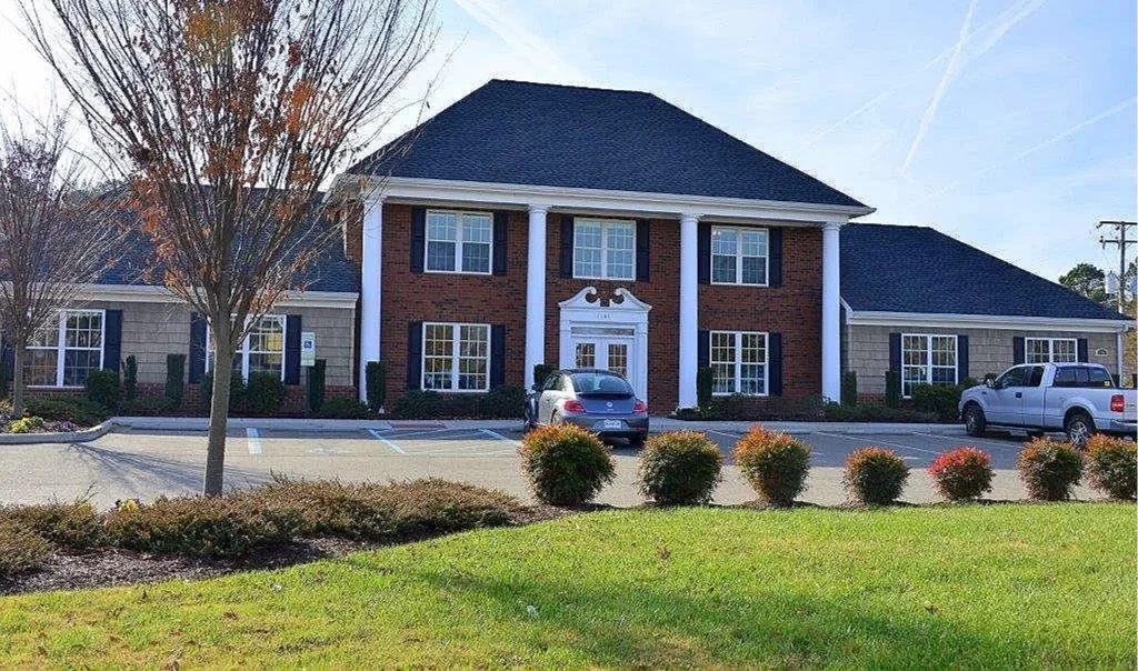 by Custom Design Works Inc. | A two-story office building with brick and siding exterior, columns at the entrance, and parking lot in front with several cars. Trees and bushes surround the building, and the sky is clear with a few contrails.