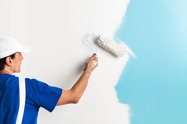 Person painting a wall with white paint using a roller, with a light blue paint section on the right.