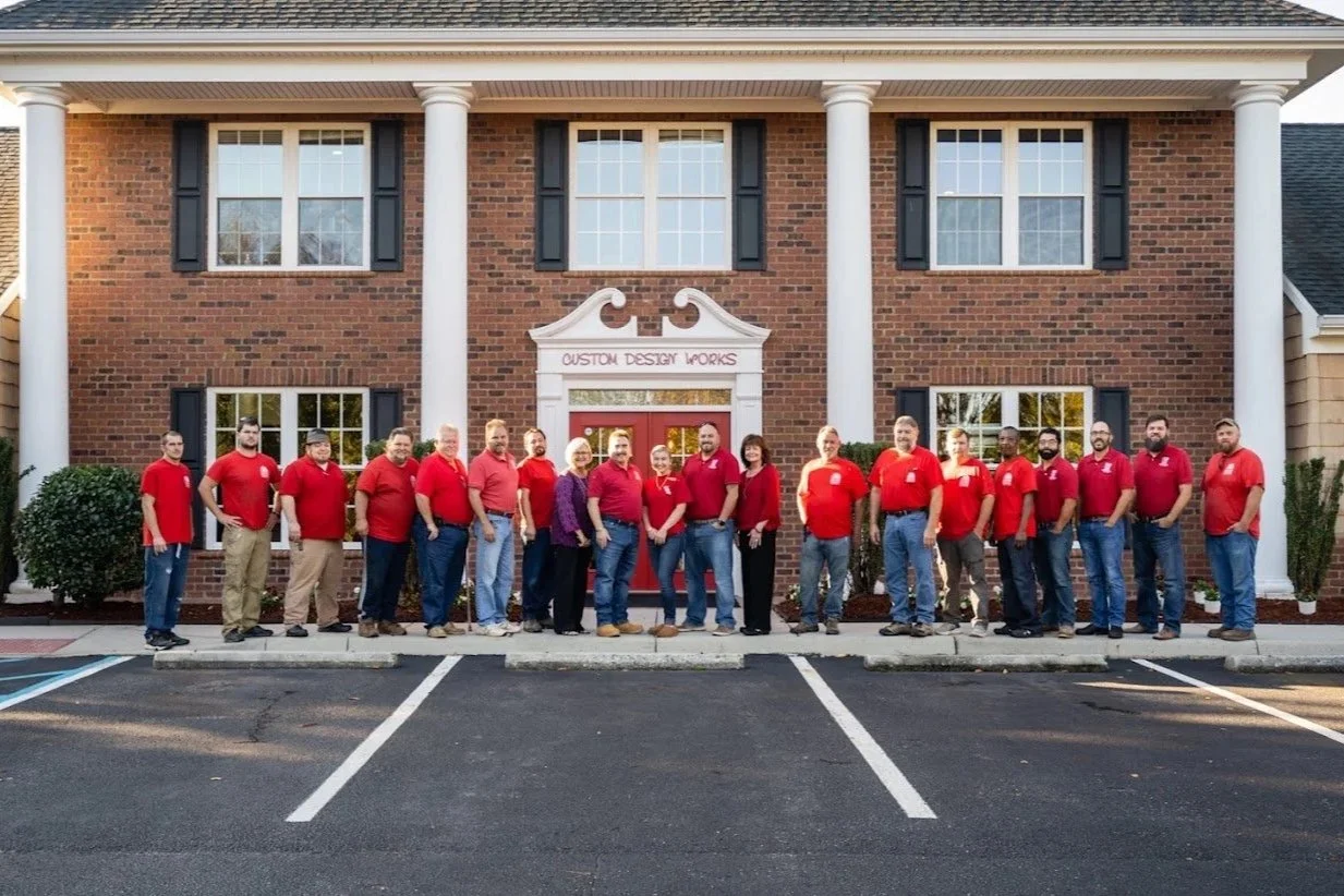 Group of people standing in front of a brick building with white columns, all wearing red shirts. The building has a sign that reads 'Custom Design Works,' with two windows and black shutters on each side.