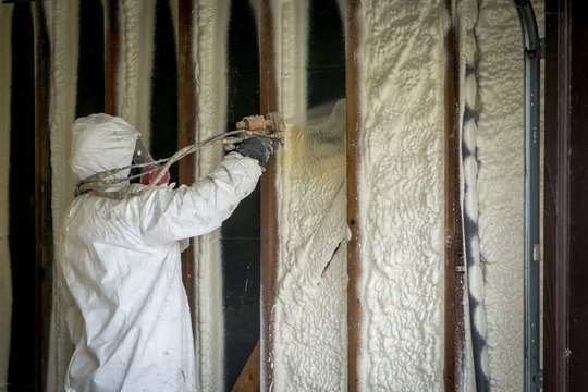 Person spraying foam insulation into wall studs inside a building.