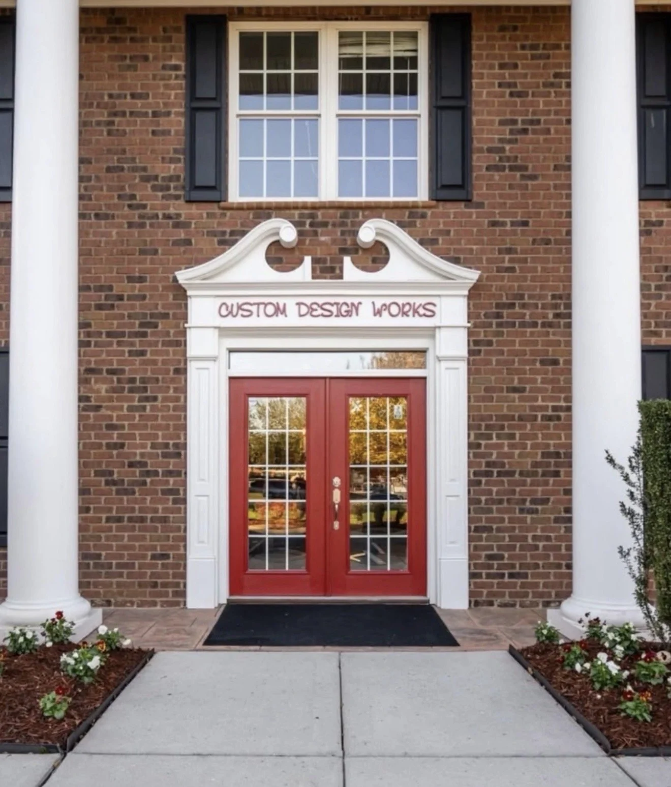 by Custom Design Works Inc. | Brick building with white columns framing a red double door entrance, a white decorative arch above the door with the sign 'Custom Design Works,' and flower beds with colorful flowers on either side of the walkway.