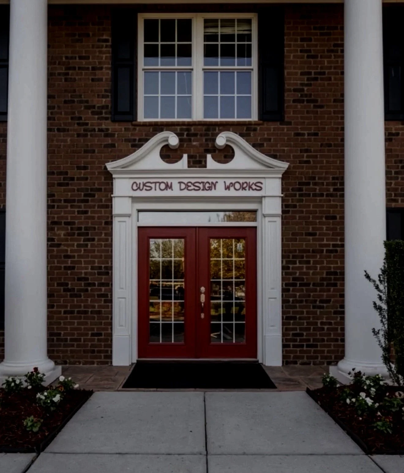 The storefront of a building with brick walls, white columns, and a red door with glass panels. Above the door, there is a sign that reads 'Custom Design Works'.
