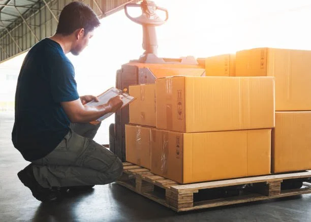 A man working in a warehouse, writing on a clipboard next to a pallet of cardboard boxes. The warehouse is well-lit with natural sunlight.