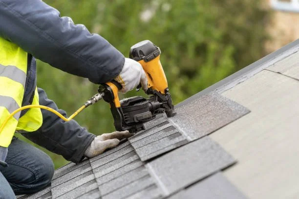 A worker in a safety vest installs roofing shingles using a nail gun on a sloped roof.