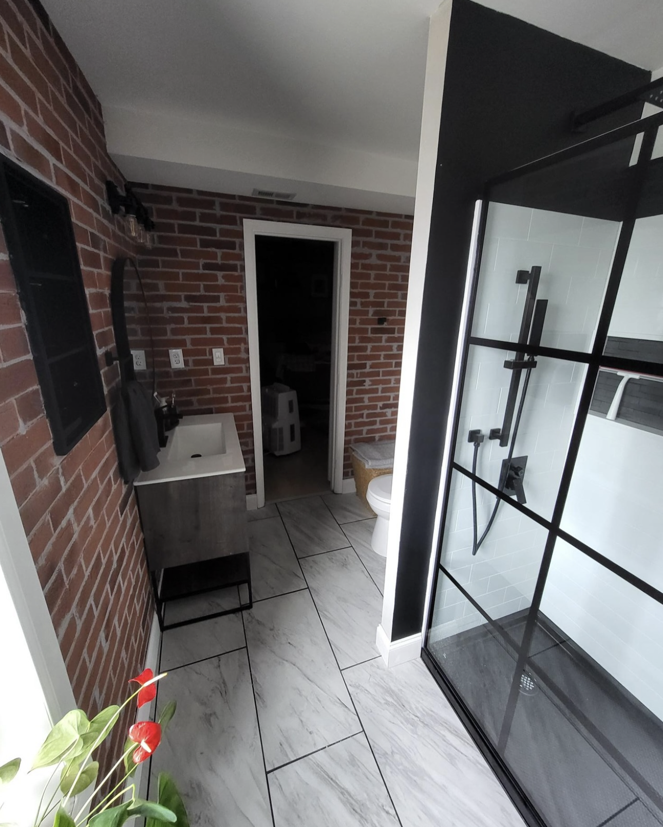 by Custom Design Works Inc. Hampton Roads construction contractor | Bathroom with brick wall, modern shower with black frame, small sink with vanity, toilet, and a laundry area in the background.