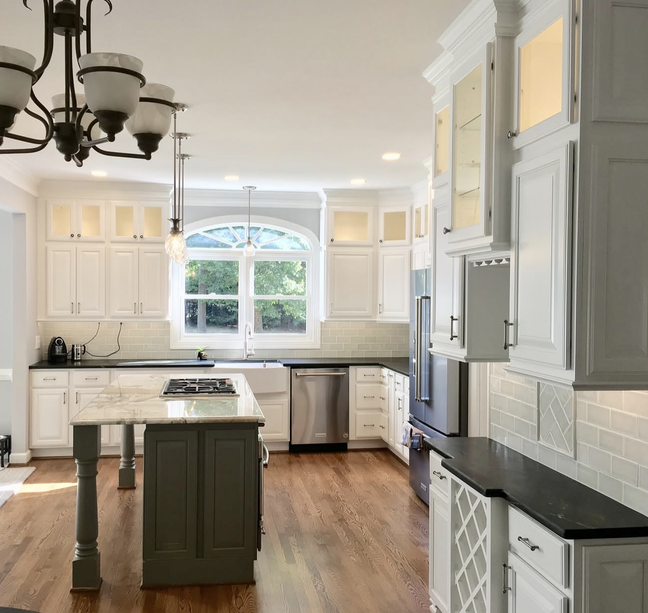 by Custom Design Works Inc. | Bright kitchen with white cabinets, black countertops, a wooden island, and a large window over the sink showing trees outside, with recessed lighting and a chandelier overhead.