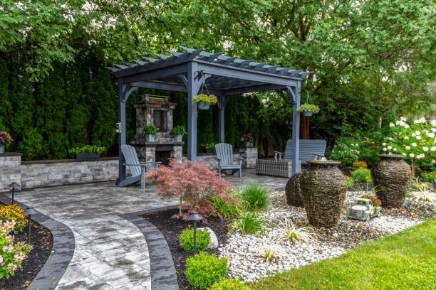 A backyard patio with a blue pergola, outdoor chairs, and decorative jars, surrounded by lush green trees and landscaping.