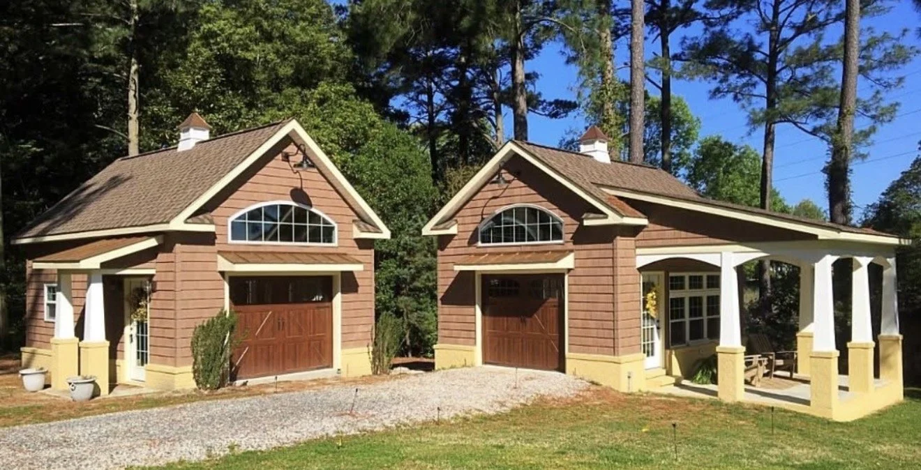 by Custom Design Works Inc. Hampton Roads construction contractor | Pair of detached garage buildings with gabled roofs, brown shingle siding, arched windows, and wooden garage doors, set against a backdrop of tall trees.