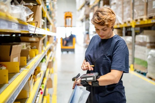 A woman working in a warehouse, wearing a navy t-shirt, using a handheld device, with shelves of boxes and inventory around her.