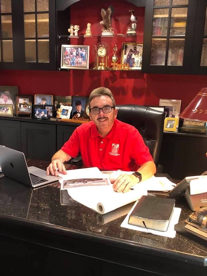 Man sitting at a computer desk in a room with framed photographs and decorative items, wearing a red polo shirt and glasses. Founder of Custom Design Works Inc.