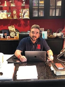 A man sitting at a cluttered desk in an office with red walls, various clocks, and decorative items behind him, using a laptop.