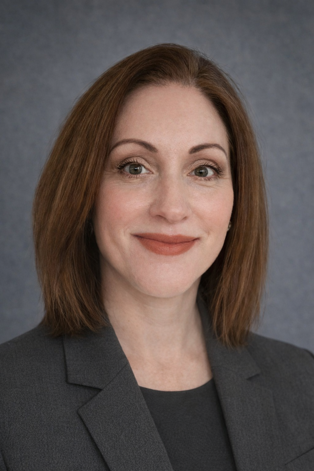 A woman with shoulder-length brown hair, wearing a dark blazer, smiling against a gray background.