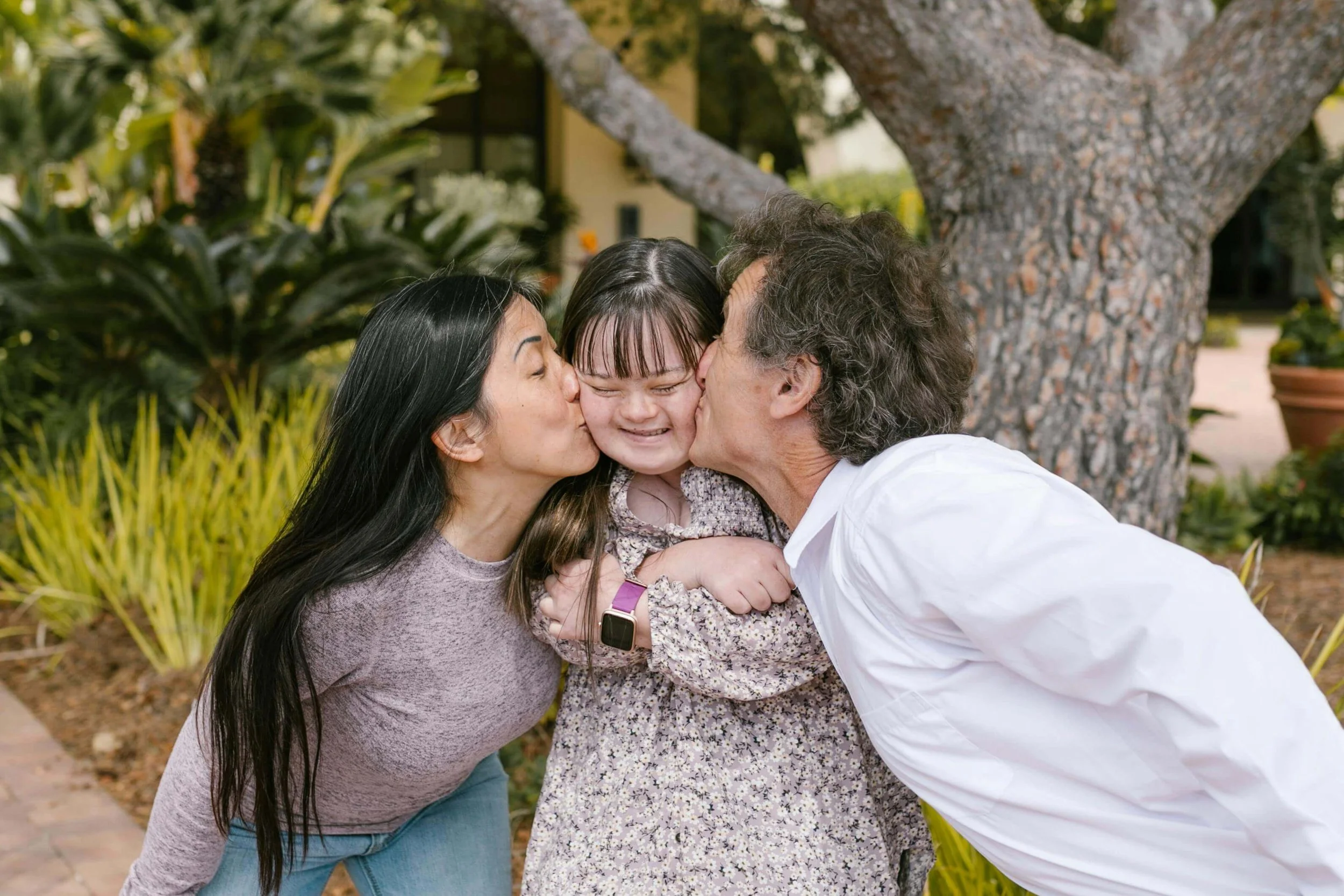 Mother and father kissing cheeks of young woman with down syndrome outside.