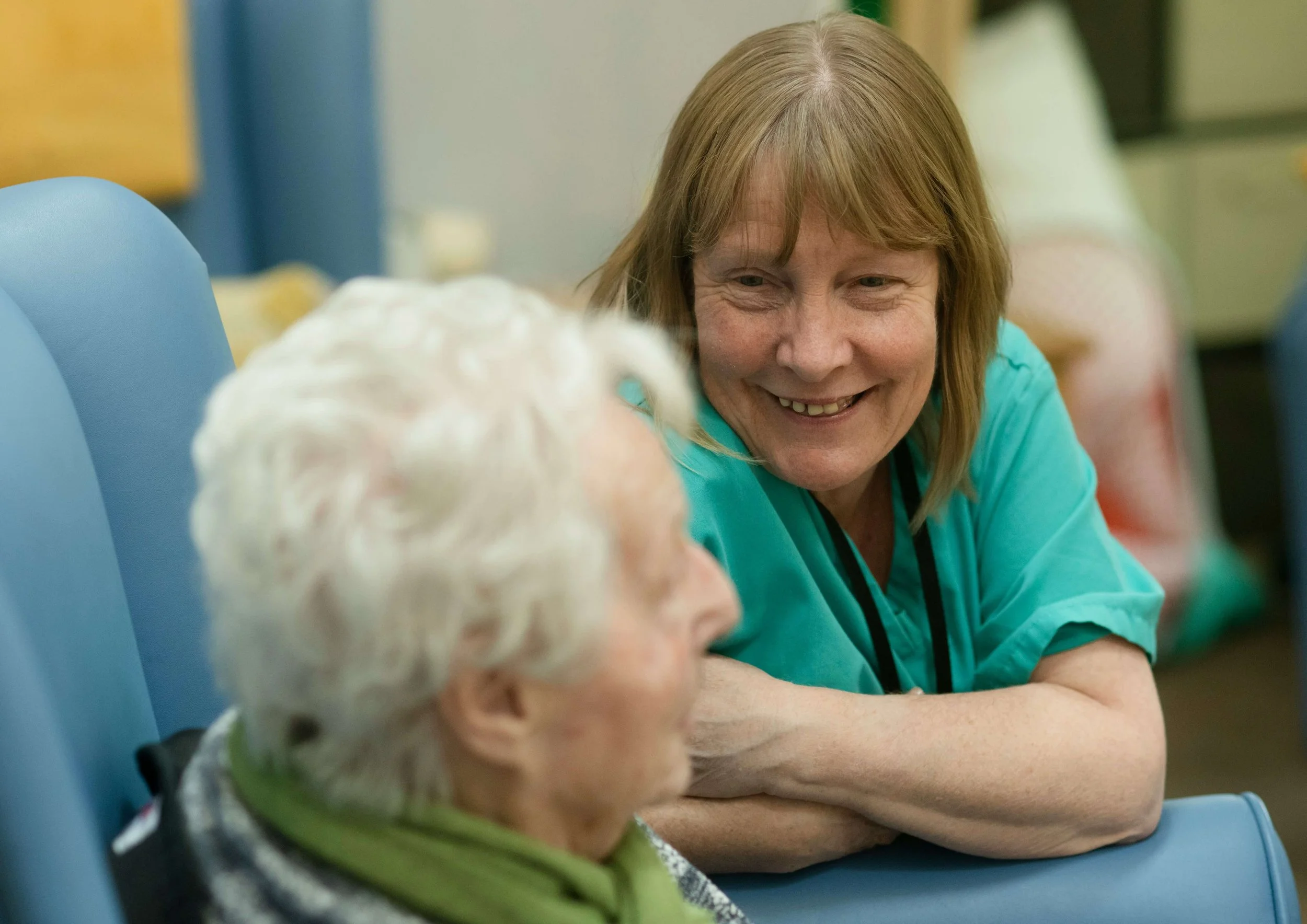 Direct support staff employee woman in scrubs smiling at elderly woman in chair in Kansas City home.