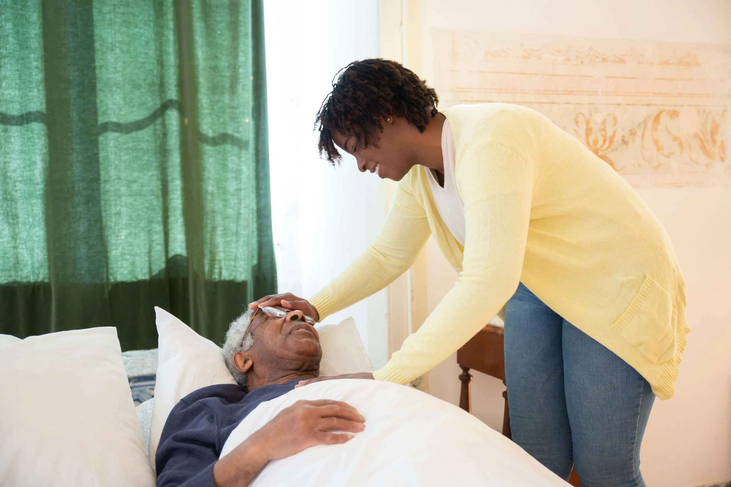 Residential health services worker smiling with hand on head of elderly man in bed at home in Kansas City.