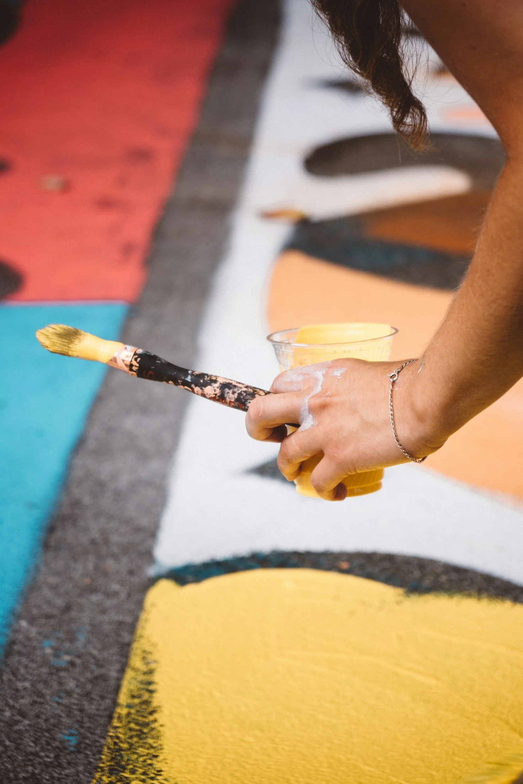Close-up of a person's hand holding a paintbrush and a small container of yellow paint, painting a colorful mural on a wall.