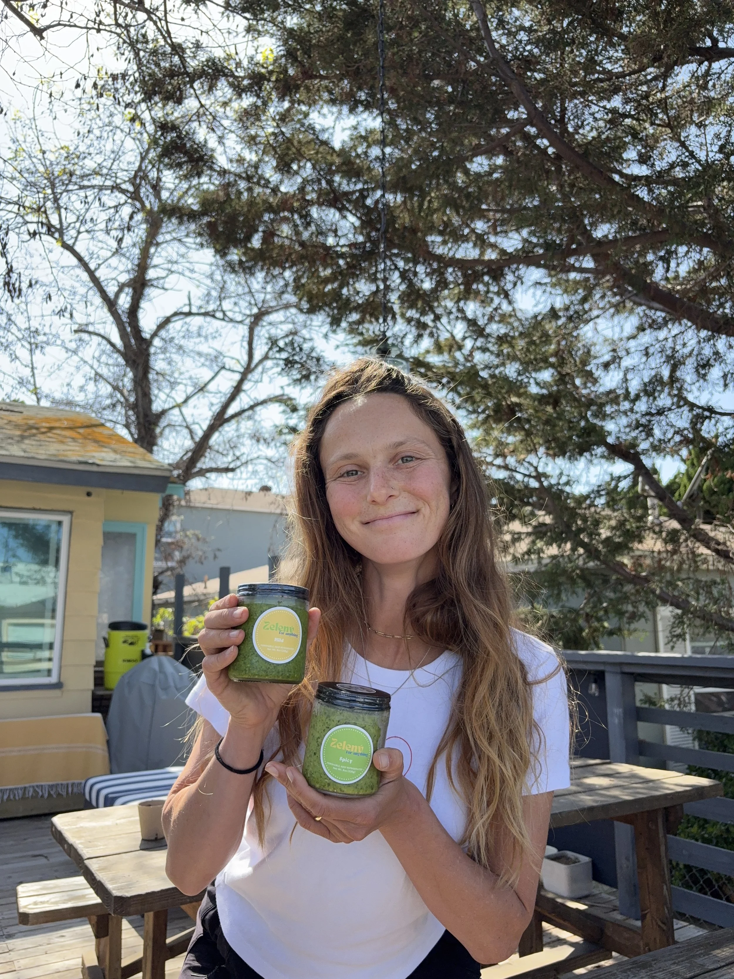 A woman with long wavy hair smiling outdoors, holding jars of green pesto sauce labeled "Zelena" and "Spicy." She is standing on a wooden deck with trees and a house in the background.