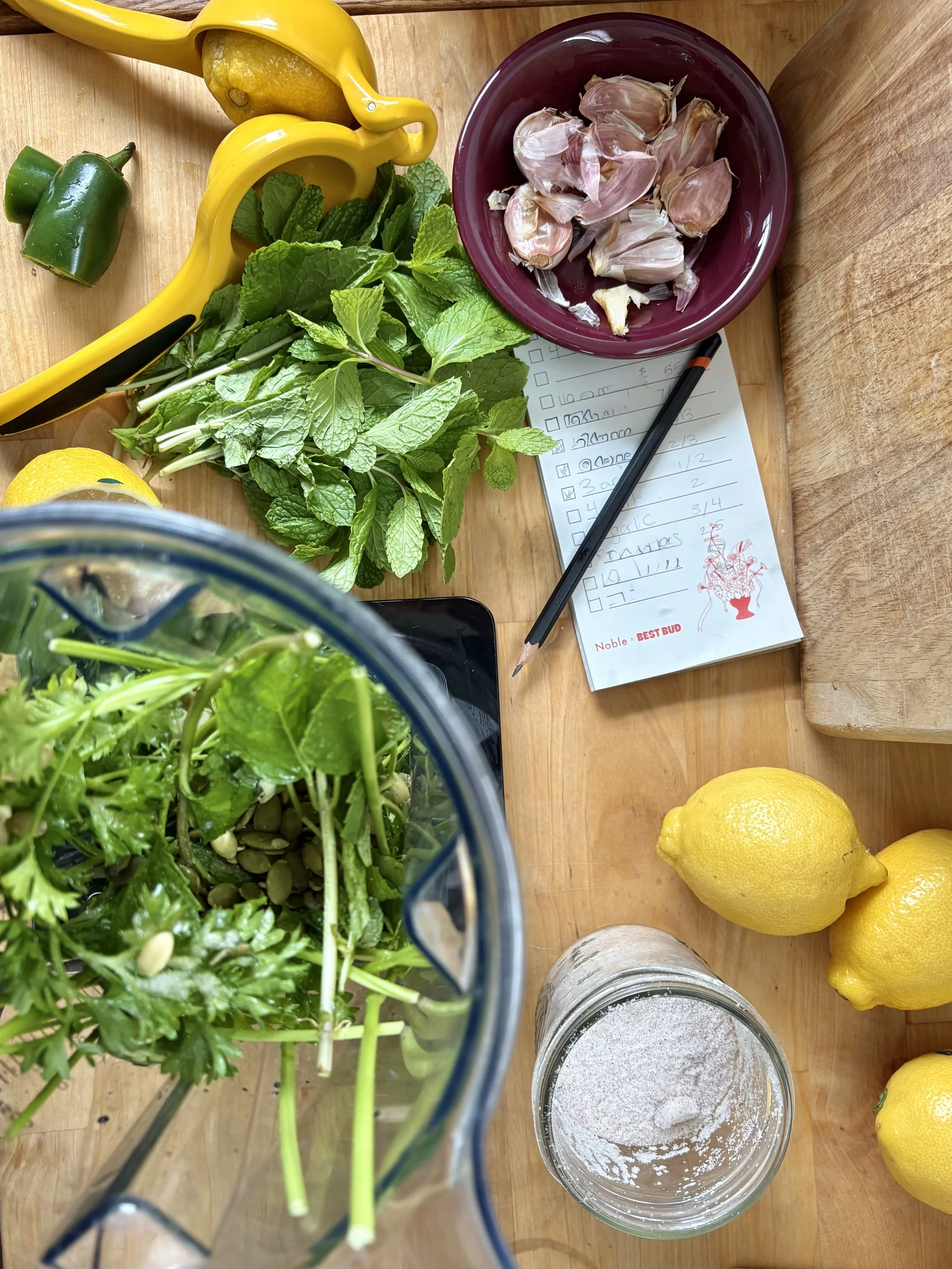 A wooden table with fresh lemons, garlic cloves in a bowl, mint and parsley herbs, green and yellow bell peppers, a glass jar with white powder, a cutting board, a notepad with handwritten notes, and a pitcher of leafy greens inside it.