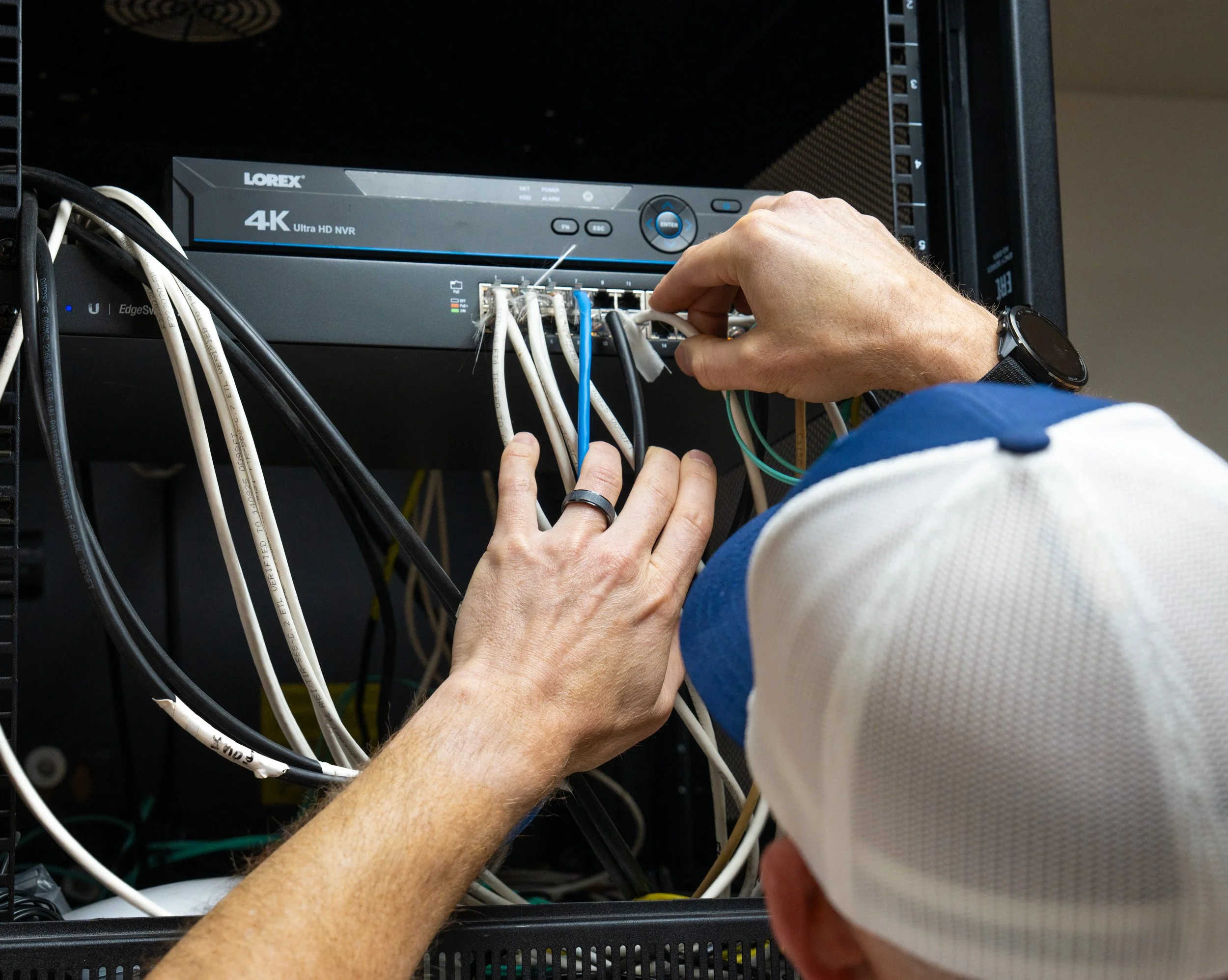 A person working on a network rack, connecting or managing cables with a hand, with a network device labeled 'Lorex 4K Ultra HD NVR' and a black server rack.
