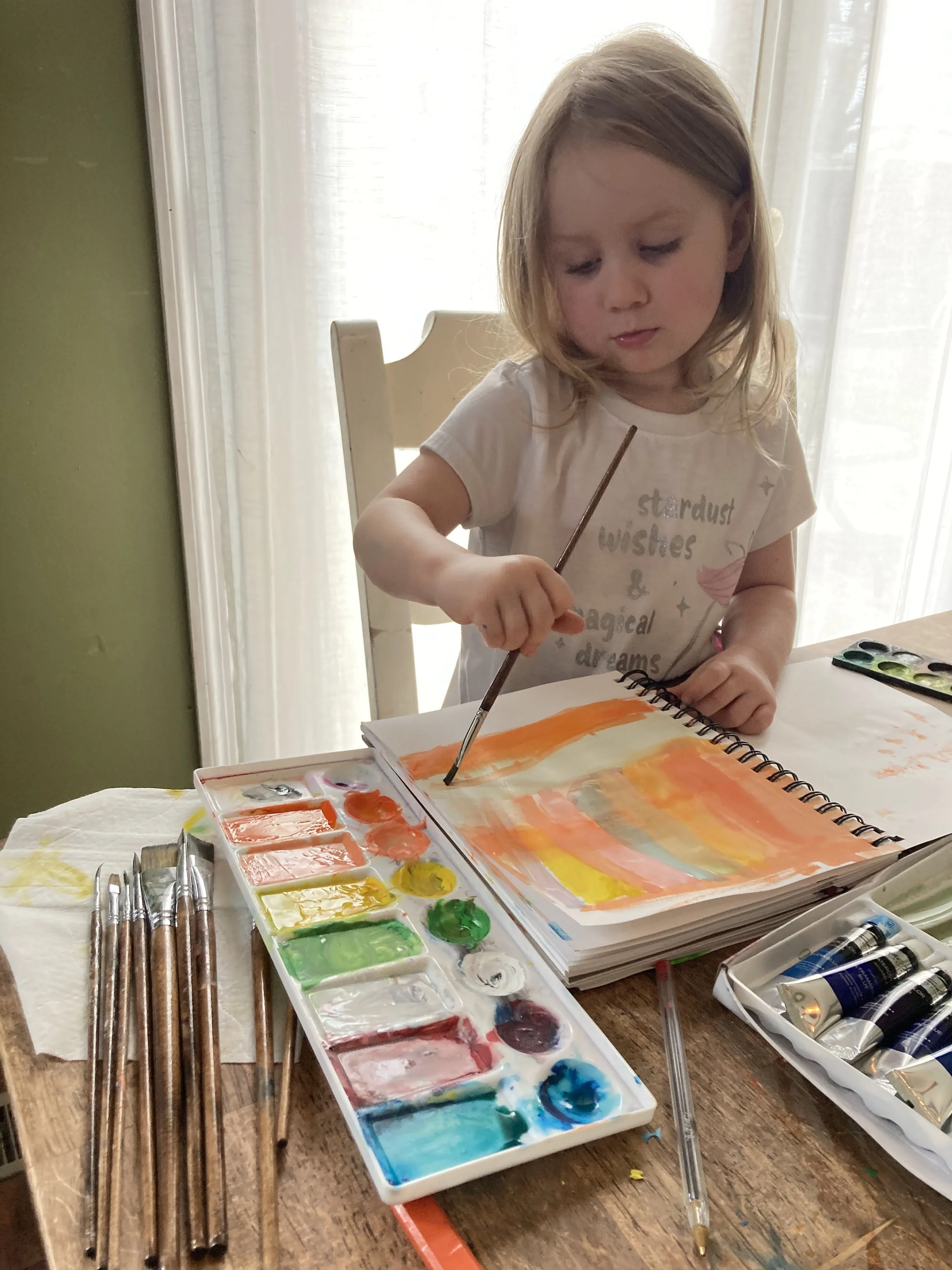 A young girl with blonde hair engaging in watercolor painting at a table, using a palette with various colors, brushes, and paper.
