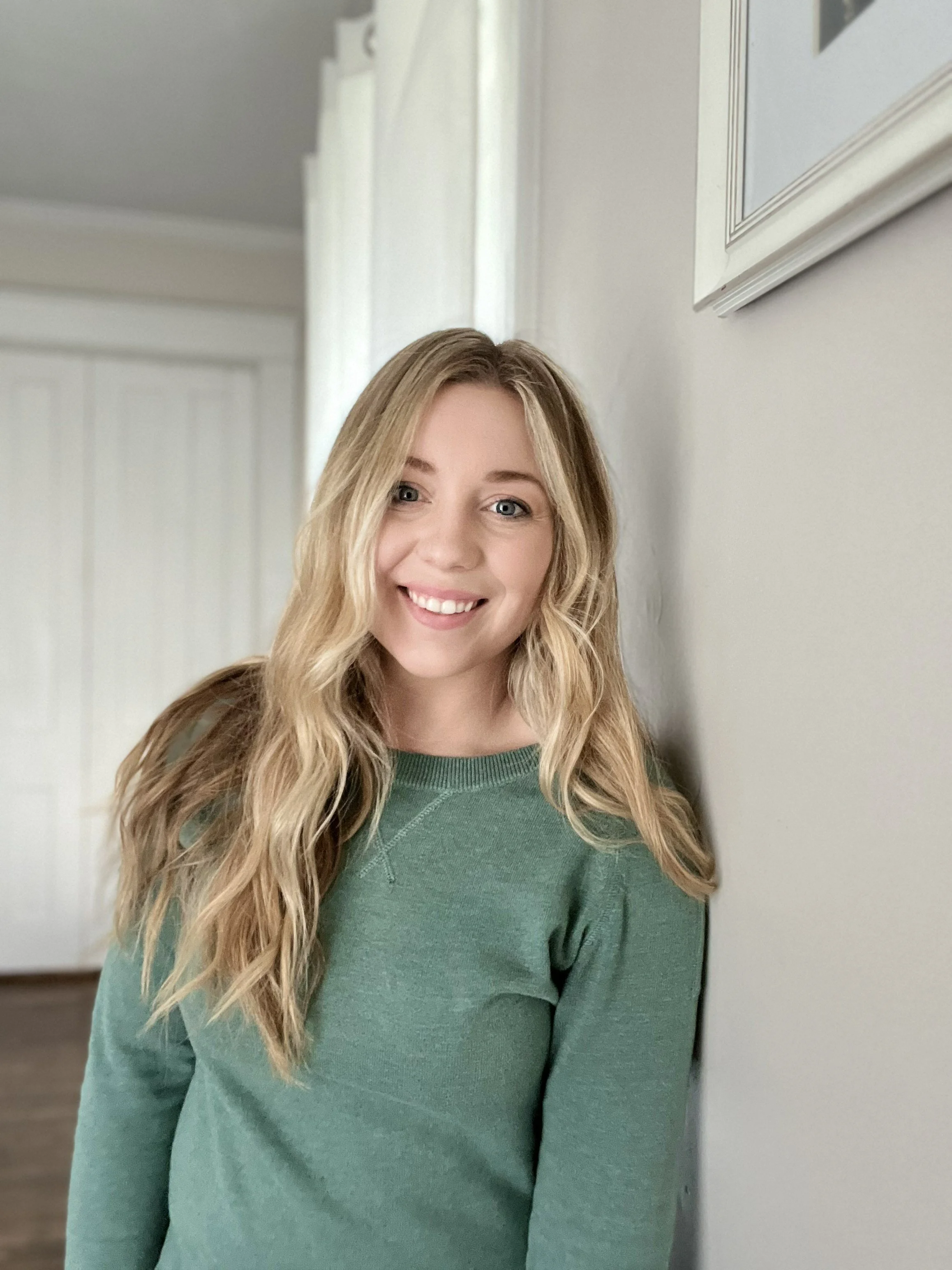 A young woman with long blonde wavy hair smiling, wearing a green sweater, standing indoors against a light-colored wall.