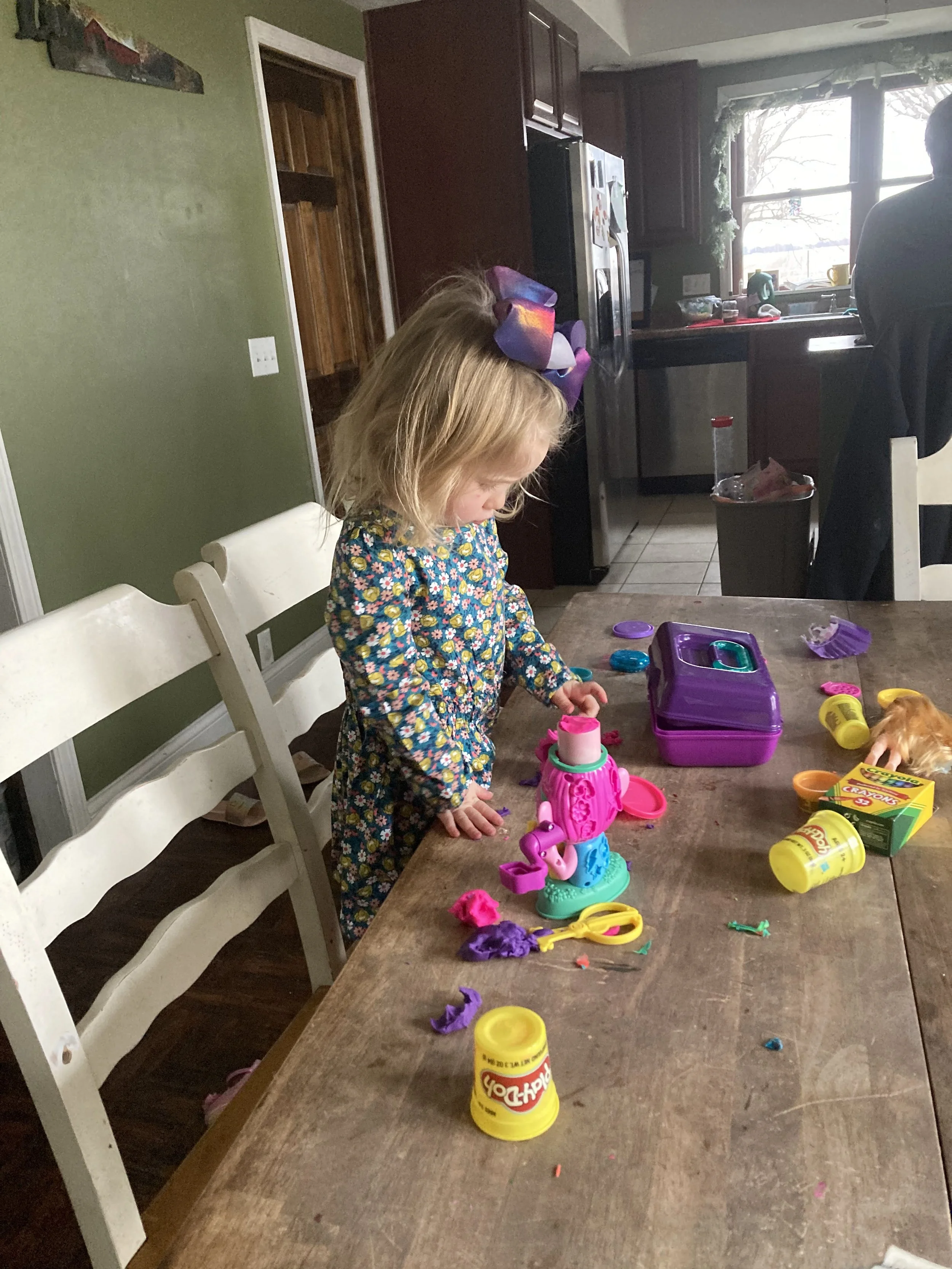 A young girl with blonde hair wearing a colorful floral dress playing with Play-Doh at a wooden table in a kitchen. The table is scattered with Play-Doh containers, molds, and tools. In the background, a man is sitting at the kitchen counter near a window, and the kitchen has dark cabinets and a window with sunlight streaming in.