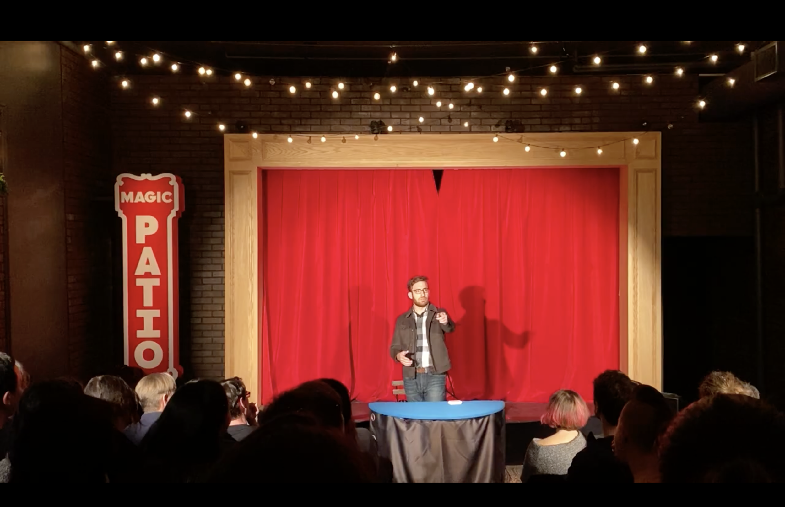 Michael Feldman performing in front of an audience at the Magic Patio in San Francisco in front of a red curtain with a yellow procenium