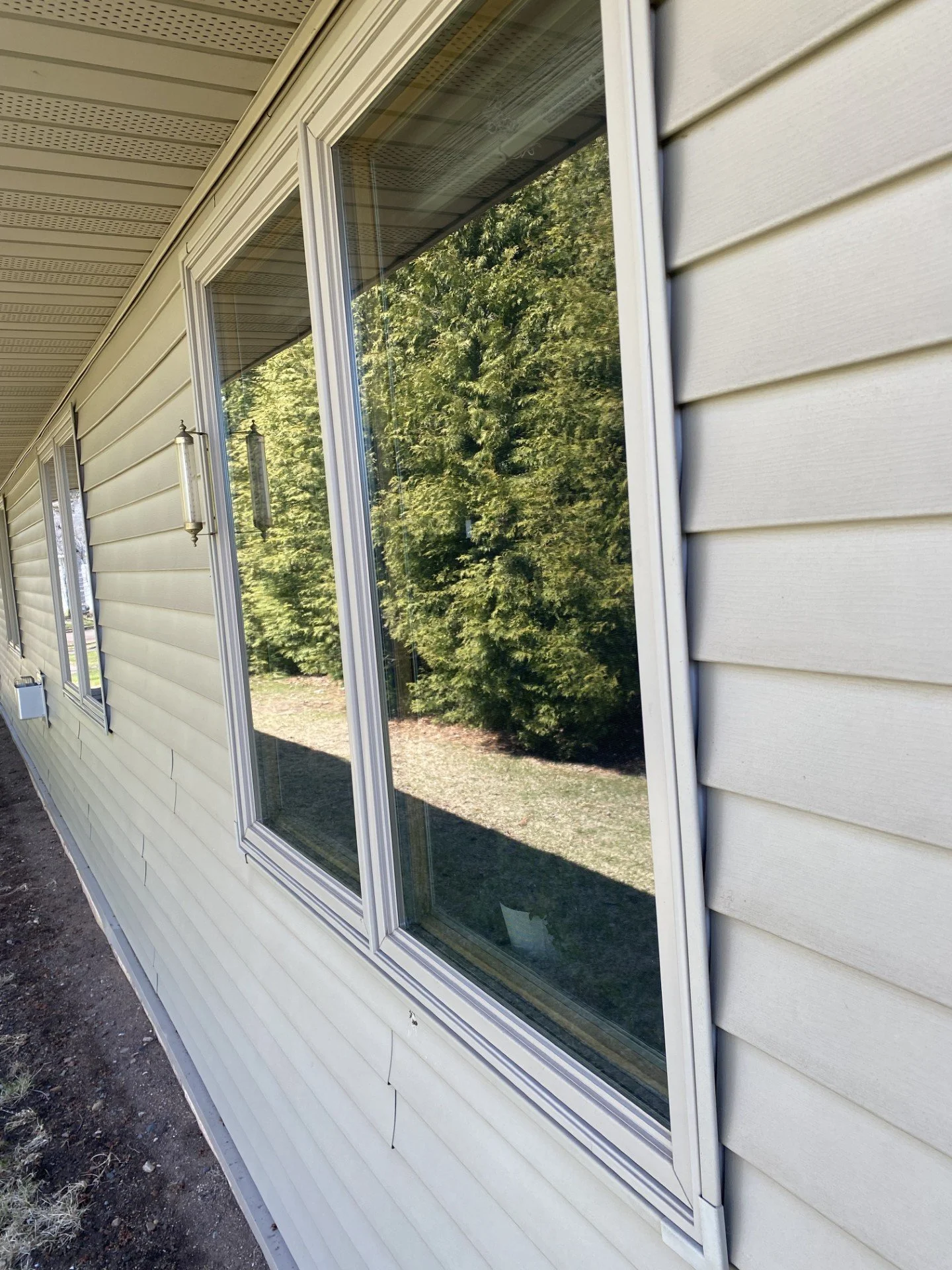 Exterior view of a house with beige vinyl siding, large window with white frame, and a reflection of trees in the glass.