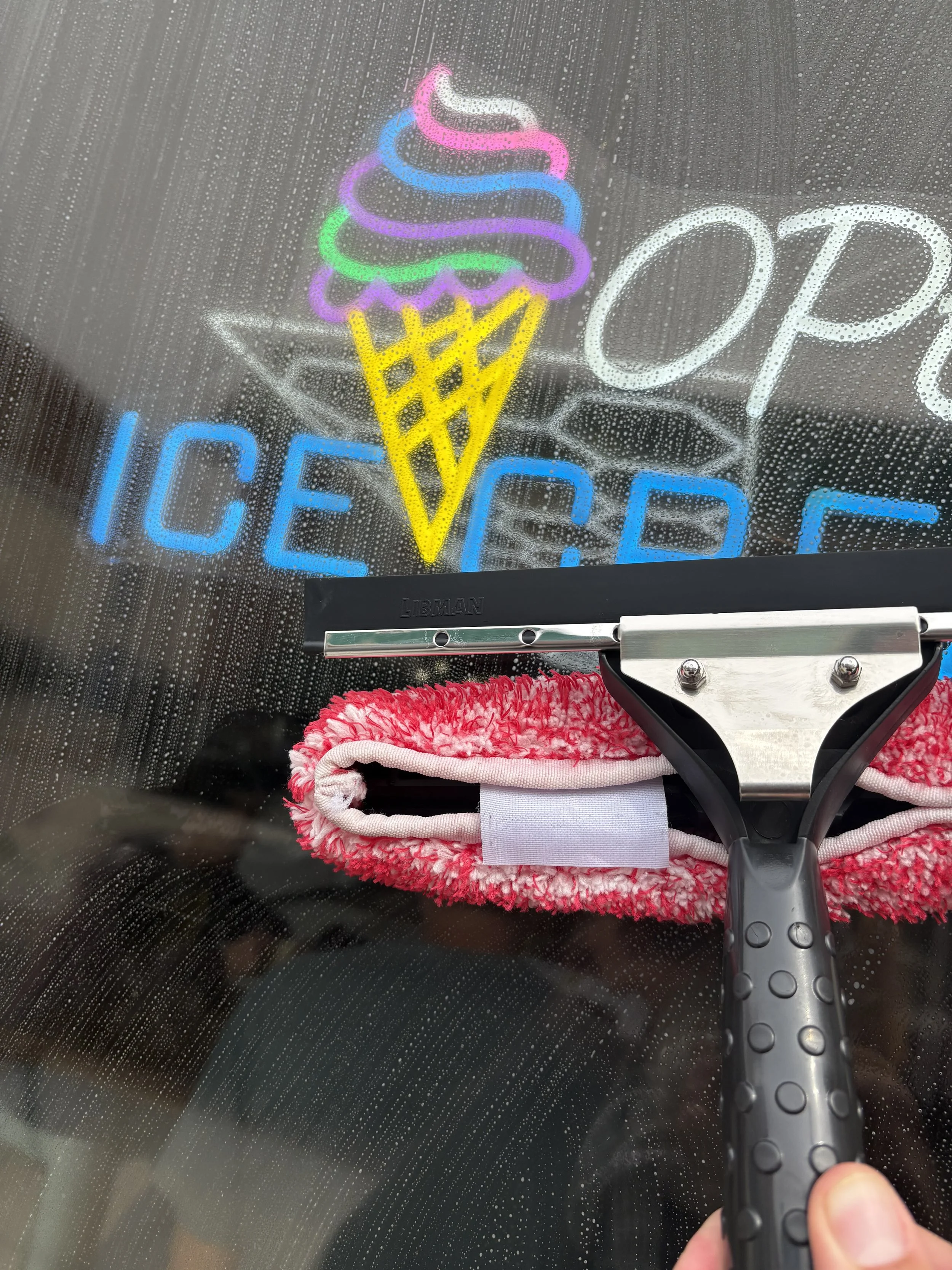 View of a squeegee cleaning a rain-covered glass window with a neon ice cream cone sign behind it, which says 'Open Ice Cream'.