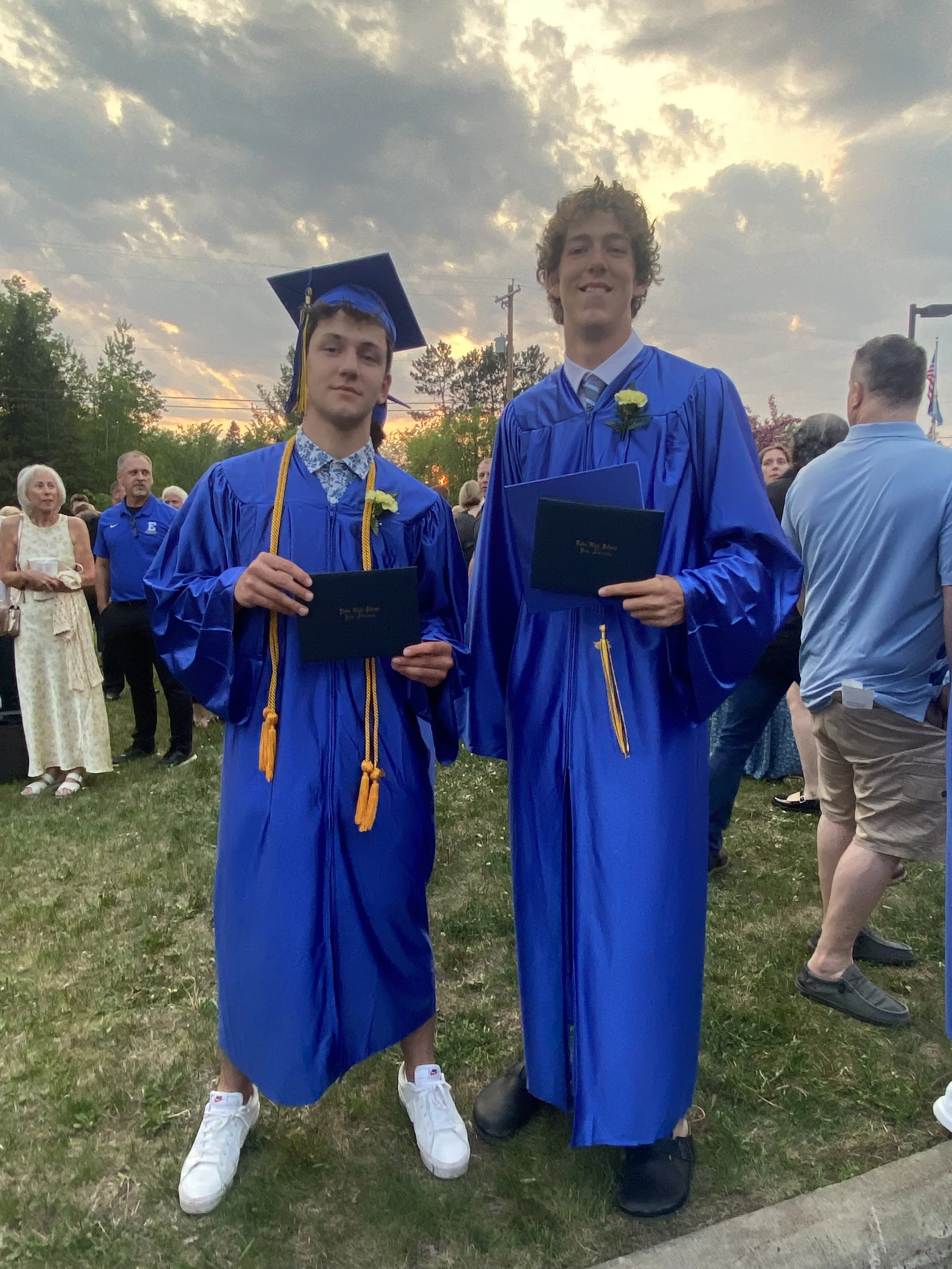 Two young men in blue graduation gowns and caps holding diplomas, standing outdoors during sunset. One student has a yellow honor cord and the other has a yellow zipper on his gown, with a sunflower on their gowns, and surrounded by other people at the graduation ceremony.