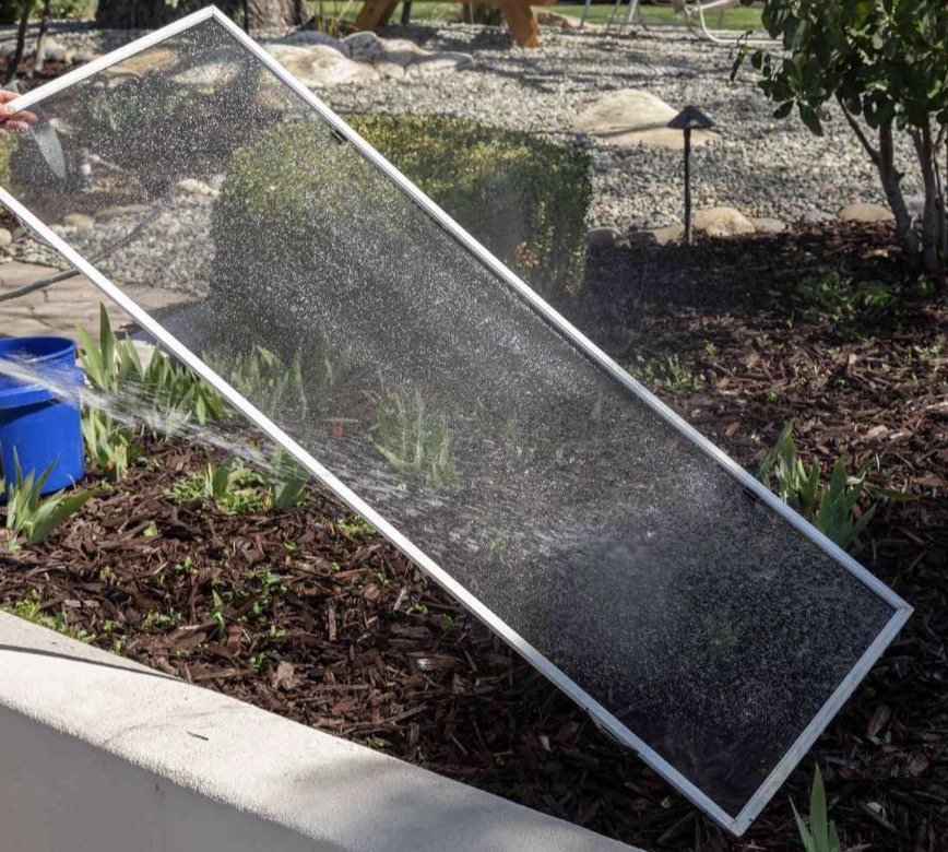 A person is cleaning a large glass window with a spray bottle and cloth in a garden with plants, mulch, and a stone path.