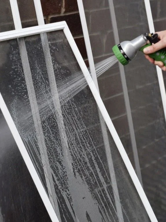 A person is spraying water on a window screen with a garden hose attachment, to clean it.