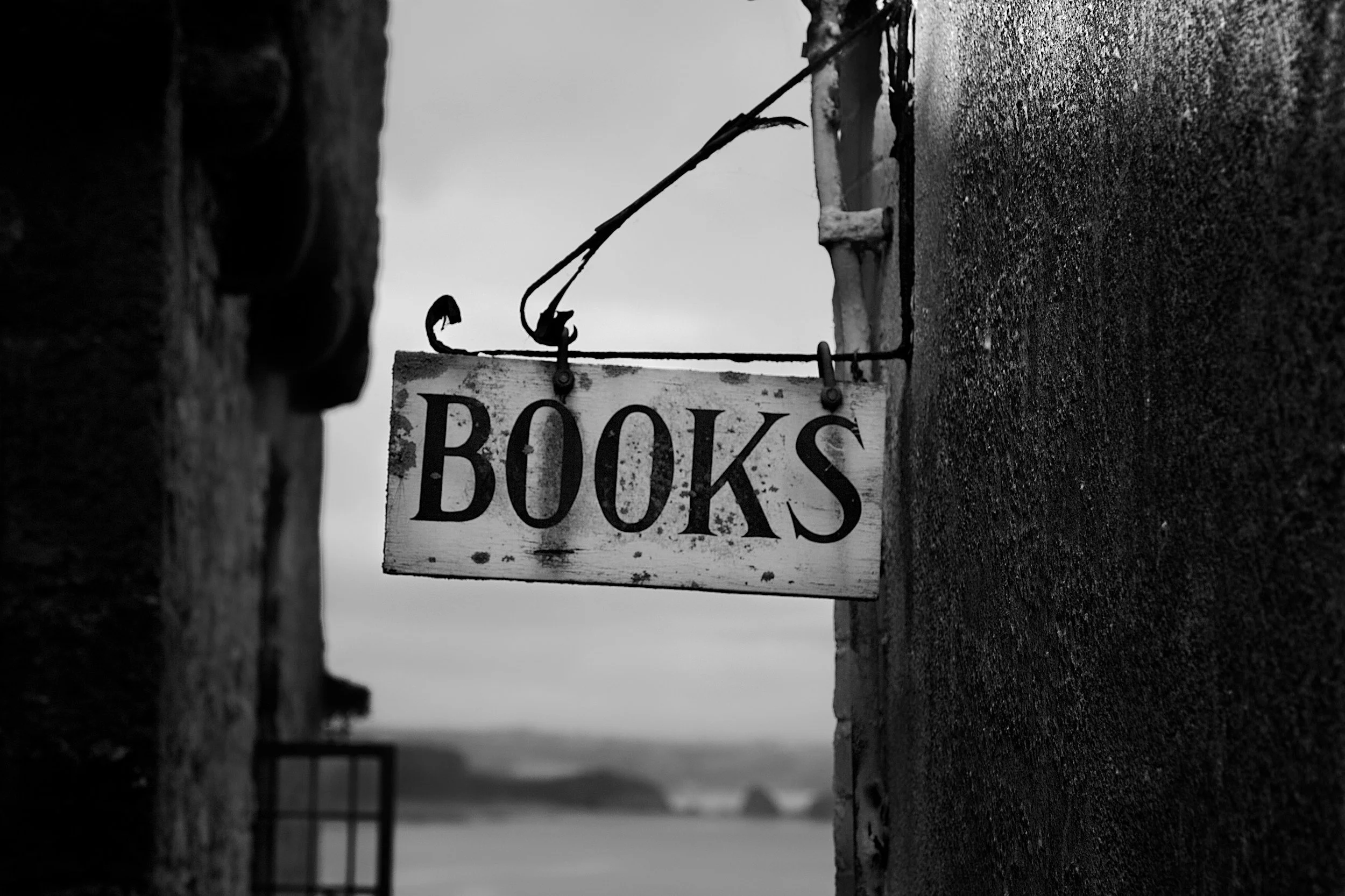 A weathered wooden sign with the word 'BOOKS' hangs between two brick walls, with a cloudy sky and distant landscape in the background.