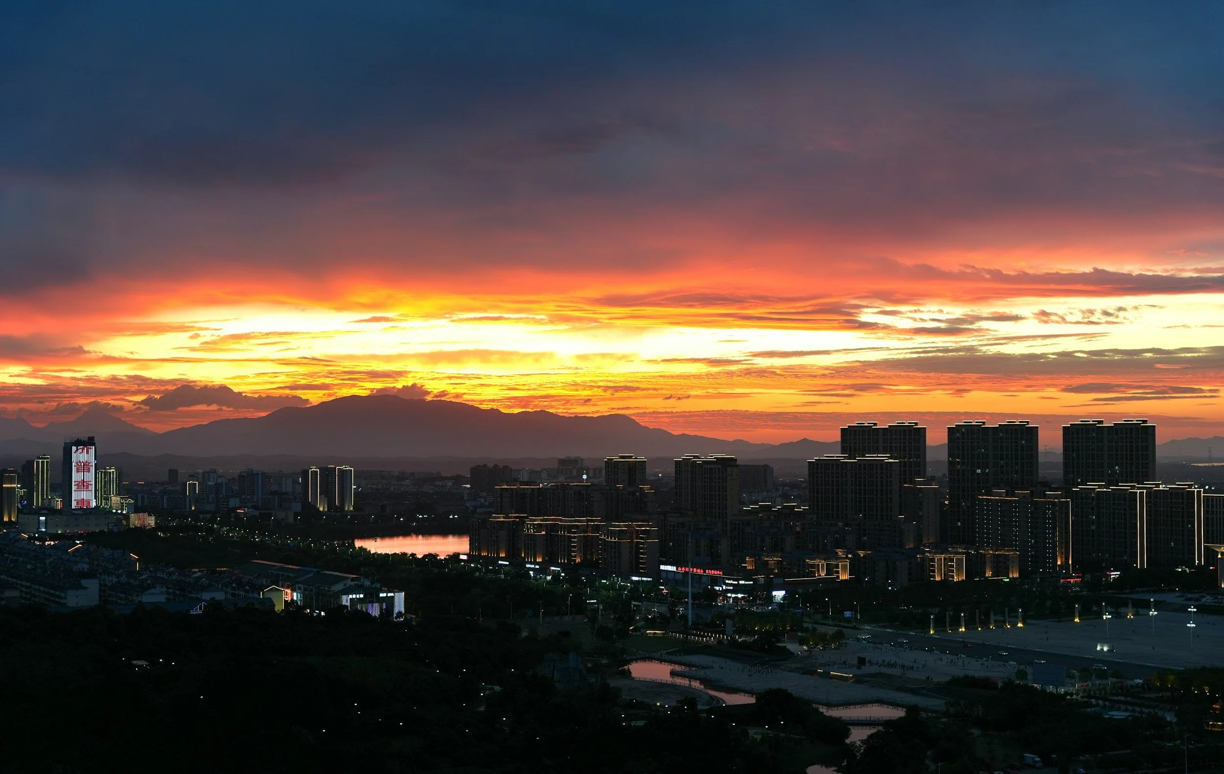 City skyline during sunset with dark clouds in the sky, mountains in the background, and illuminated buildings in the foreground.