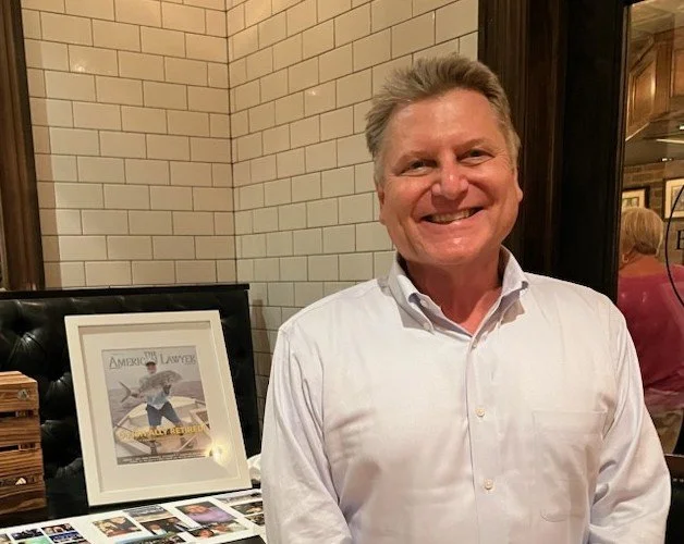 Allan Milloy in a white shirt standing indoors near framed artwork and photographs on a table.