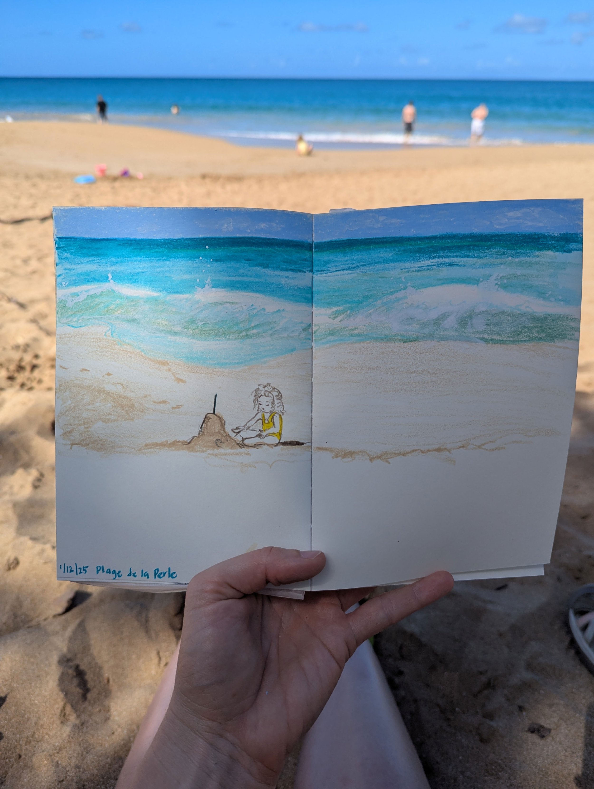 Photo of a hand holding a sketchbook at the beach, showing a watercolor painting of the ocean and a girl building a sandcastle, with people in the background on the sandy beach and water.