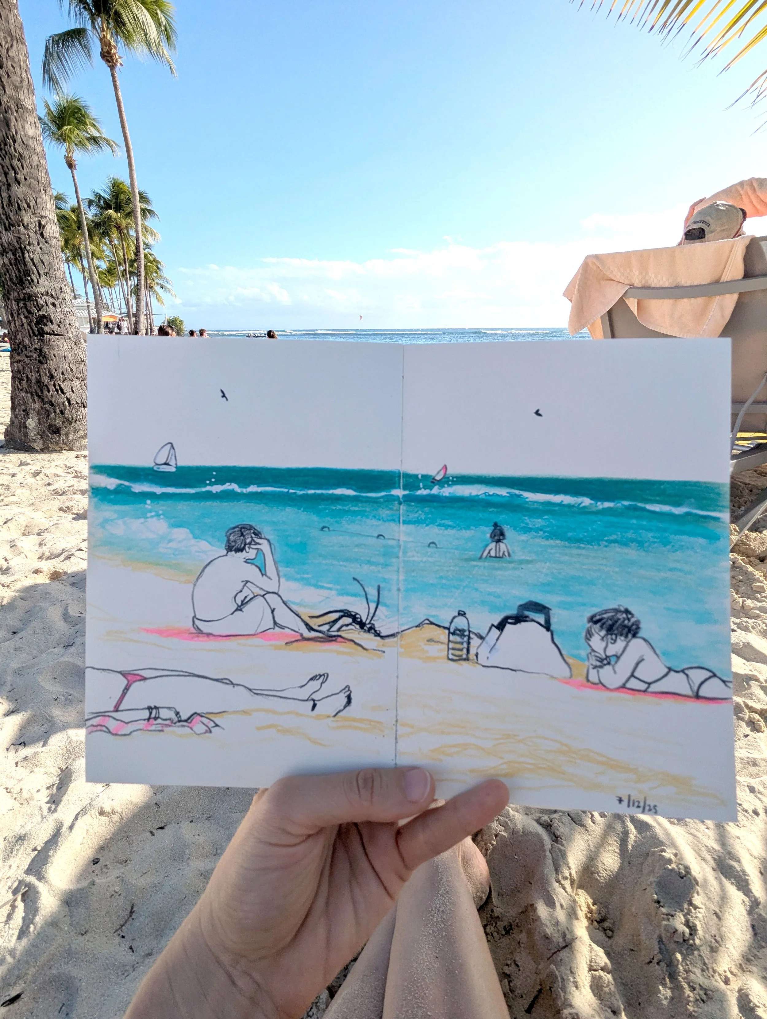 A person holding a sketchbook with a drawing of people relaxing on a beach near palm trees, with the real beach and ocean in the background.