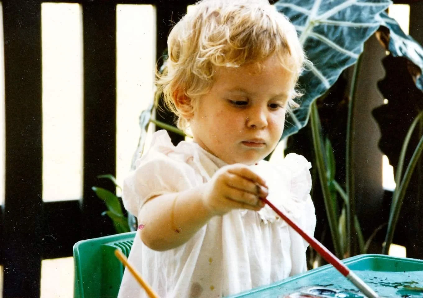 Young girl with curly blonde hair paints using a red brush. She wears a white shirt and is seated at a table with a teal tray, surrounded by green plants and a black railing.