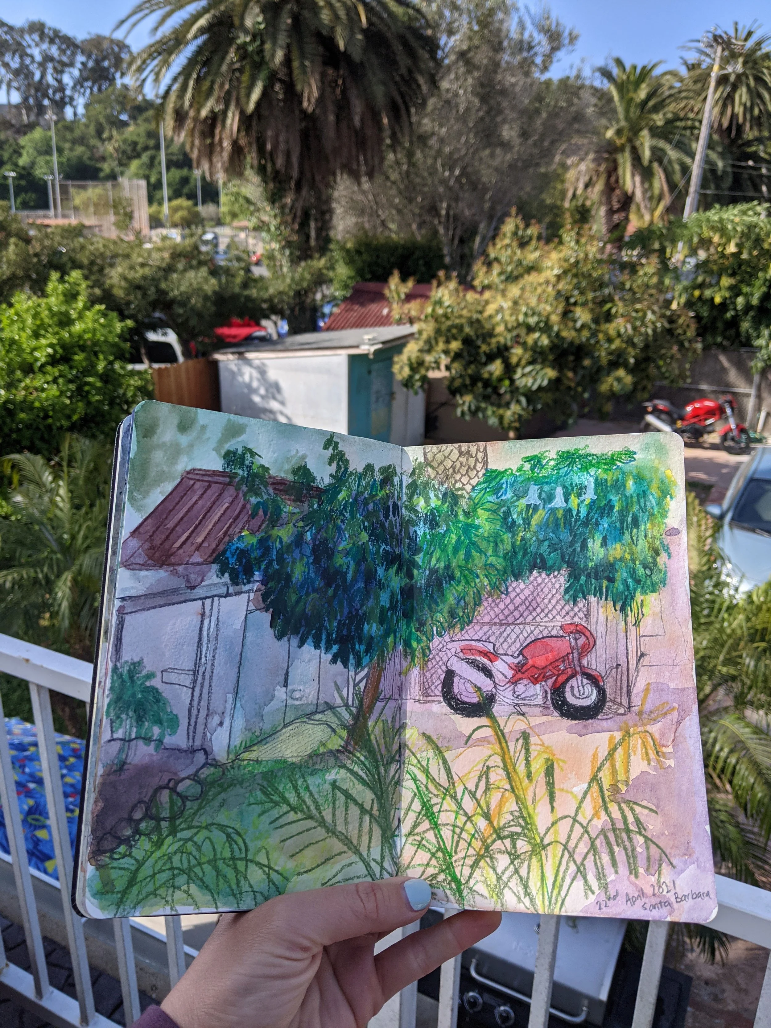 A hand holding a sketchbook with a colorful drawing of a backyard scene, including trees, a red motorcycle, and a small shed, against a background of lush greenery and partly cloudy sky.
