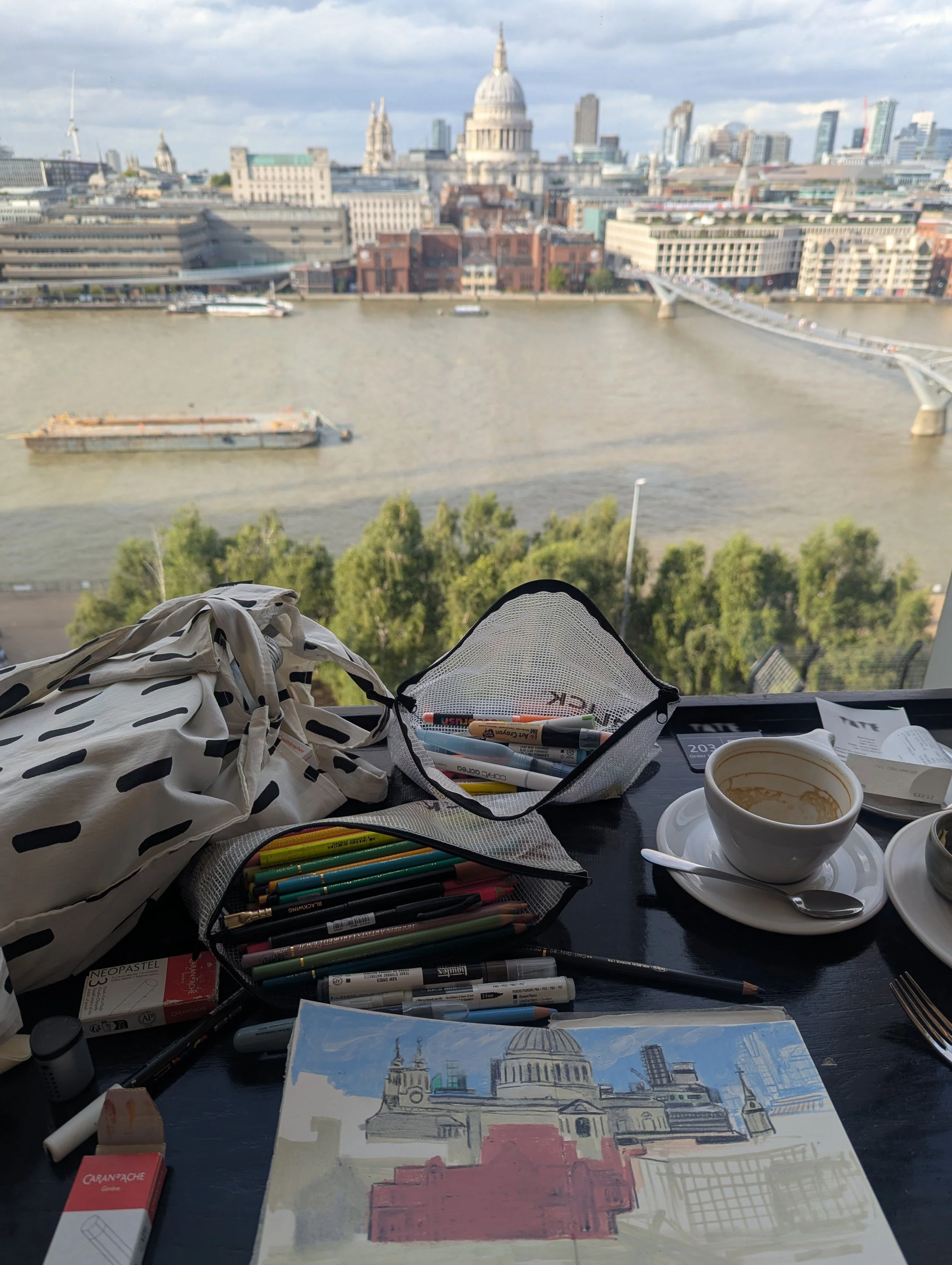 View of the London skyline including St. Paul's Cathedral, from a table with art supplies, a coffee cup, and a sketch of the city.