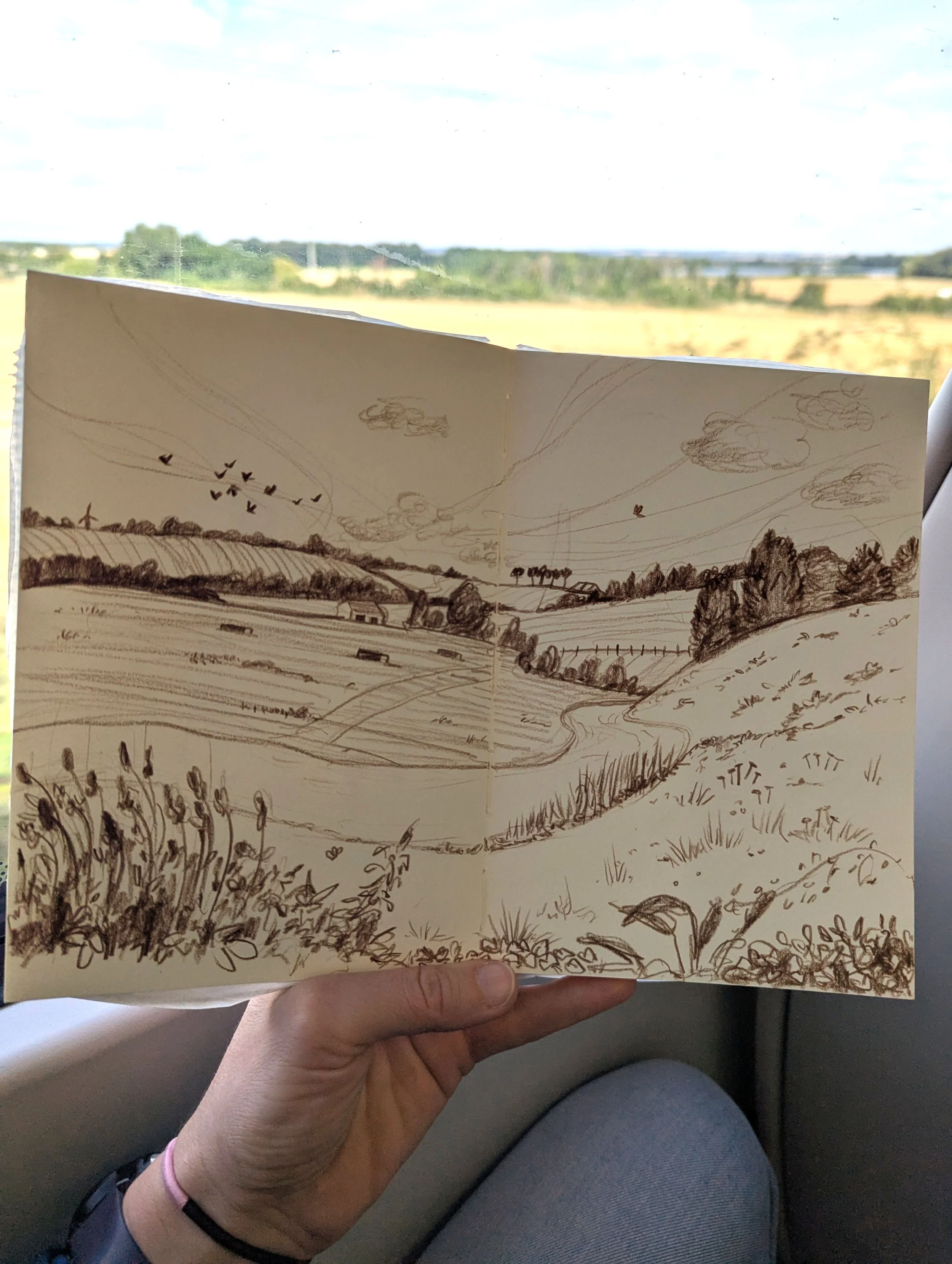 A hand holding a sketchbook with a landscape drawing of rolling hills, trees, and a few clouds, with a farm scene, in front of a train window showing an open countryside with fields and trees.