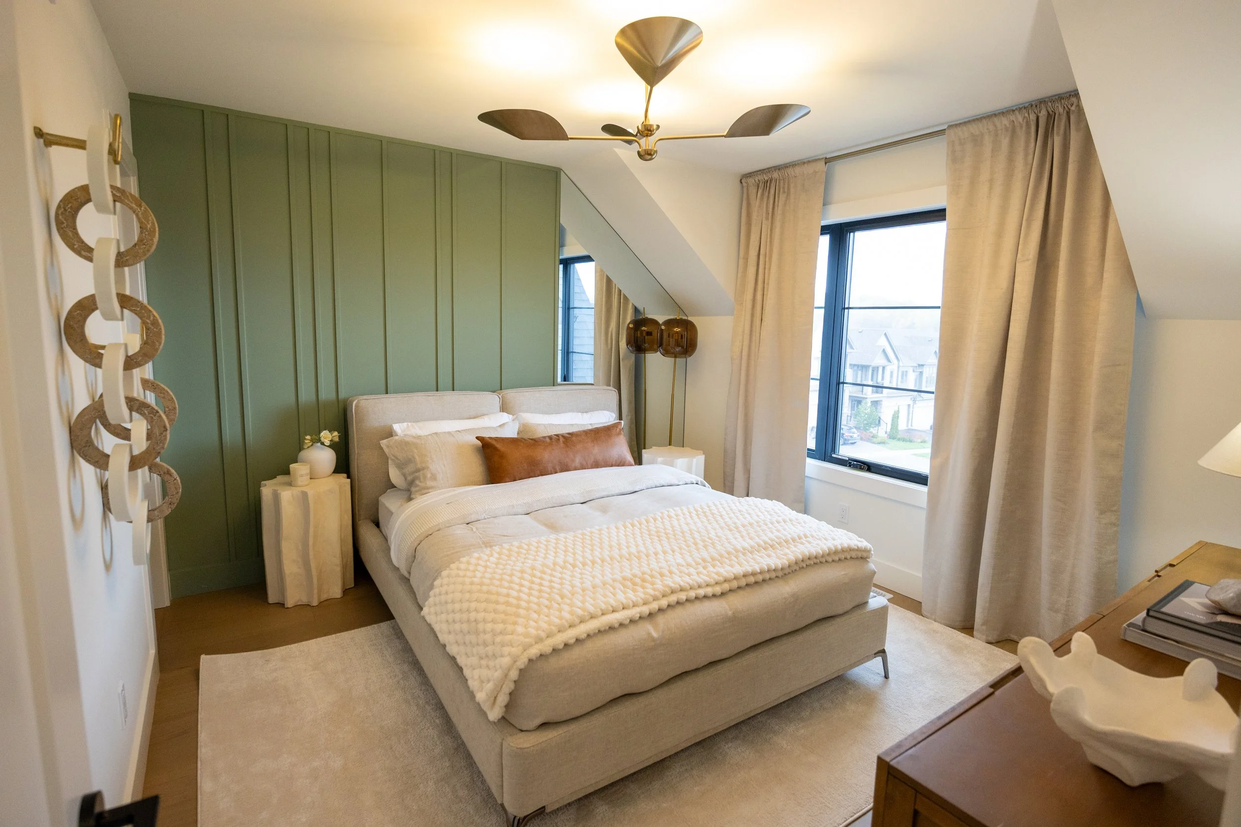 A bedroom with a beige upholstered bed, cream bedding, and a brown pillow, set against a green paneled accent wall. There are two hanging pendant lights and large windows with beige curtains. A ceiling fan with wooden blades and a brass finish is mou