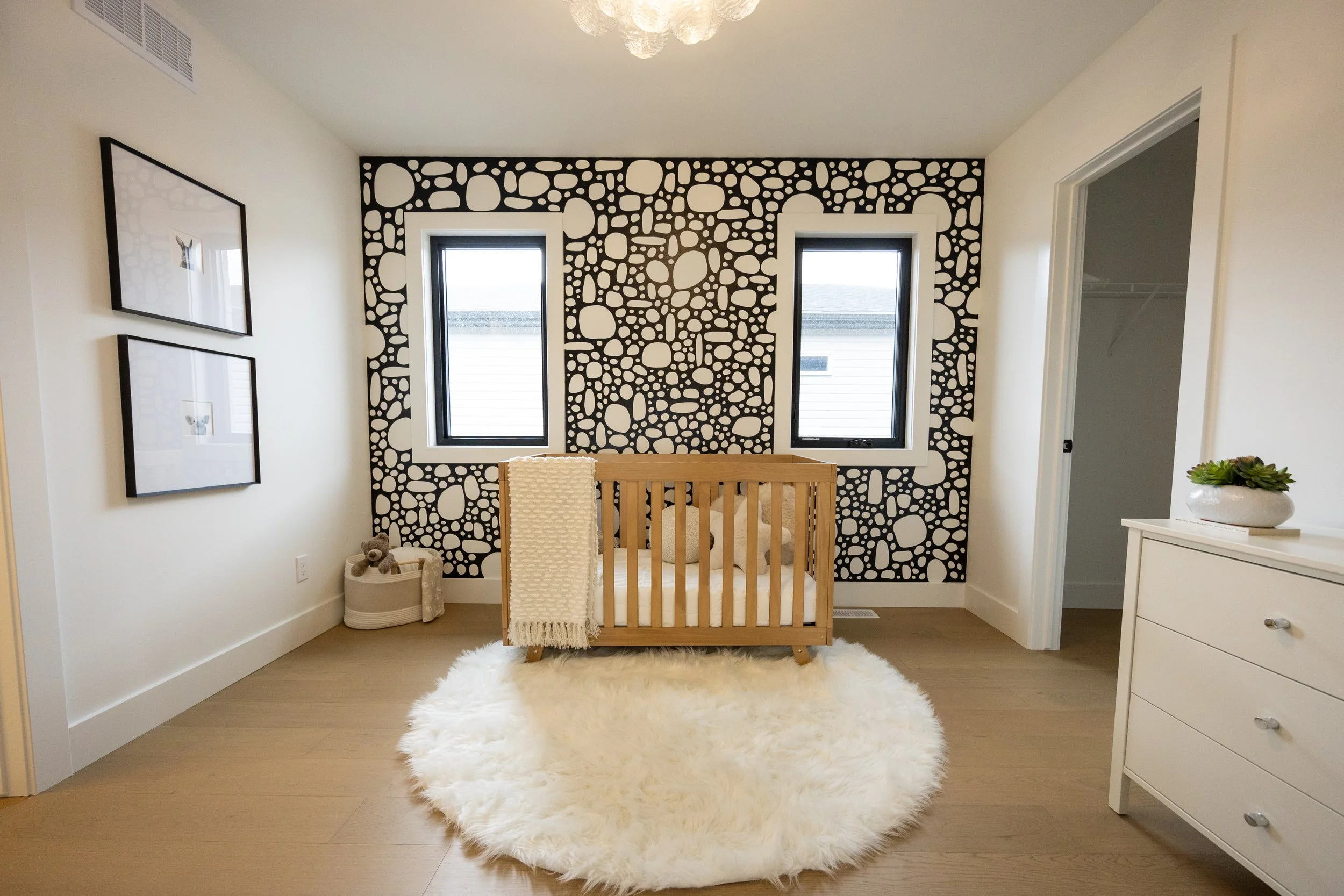 A cozy nursery with a white fluffy rug, a wooden crib with a beige blanket, and a black and white polka dot accent wall with two windows.