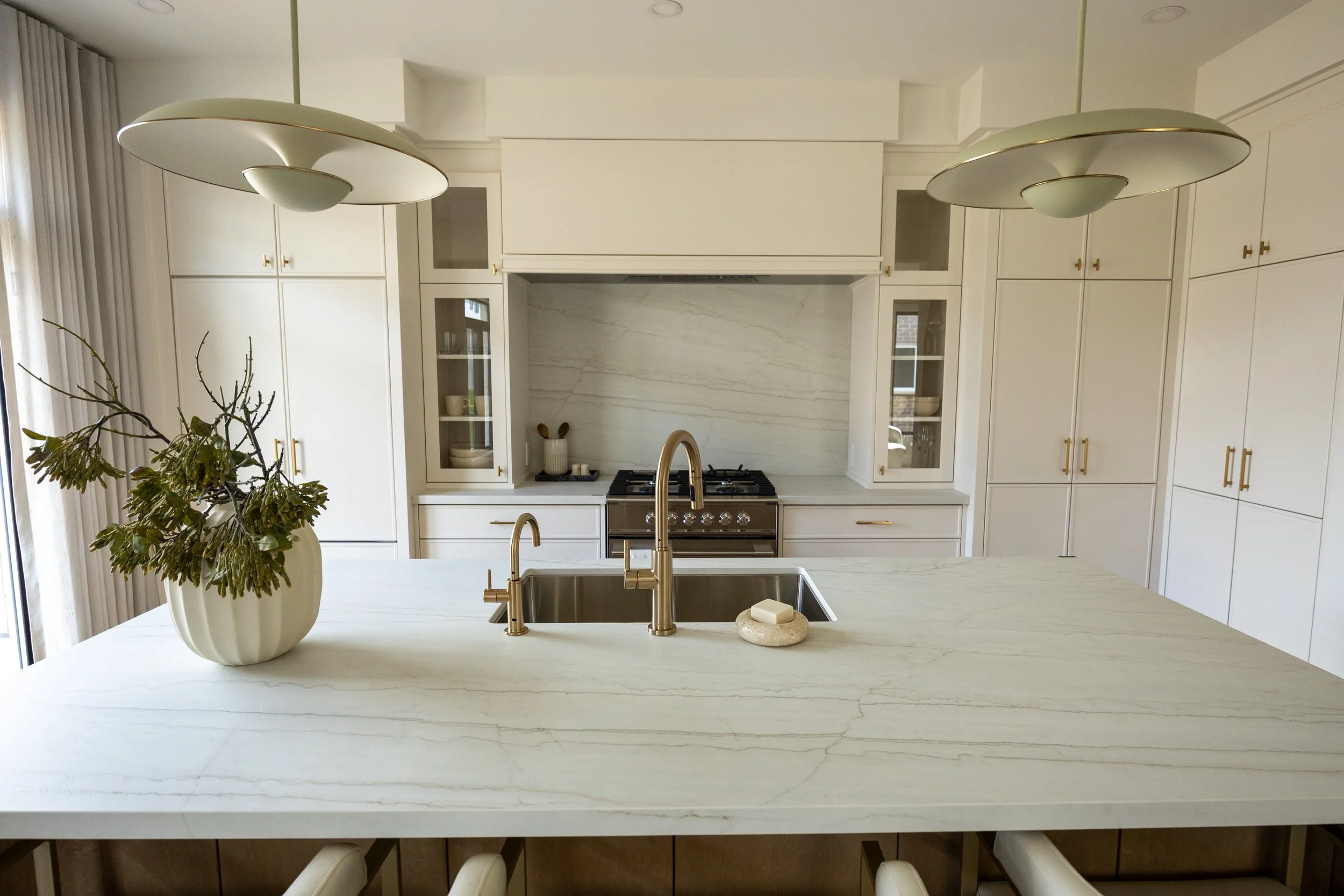 Modern kitchen with white cabinets, marble backsplash, and a white countertop. A gold faucet is at the sink, and there is a vase with green foliage on the counter. Two stylish pendant lights hang from the ceiling.