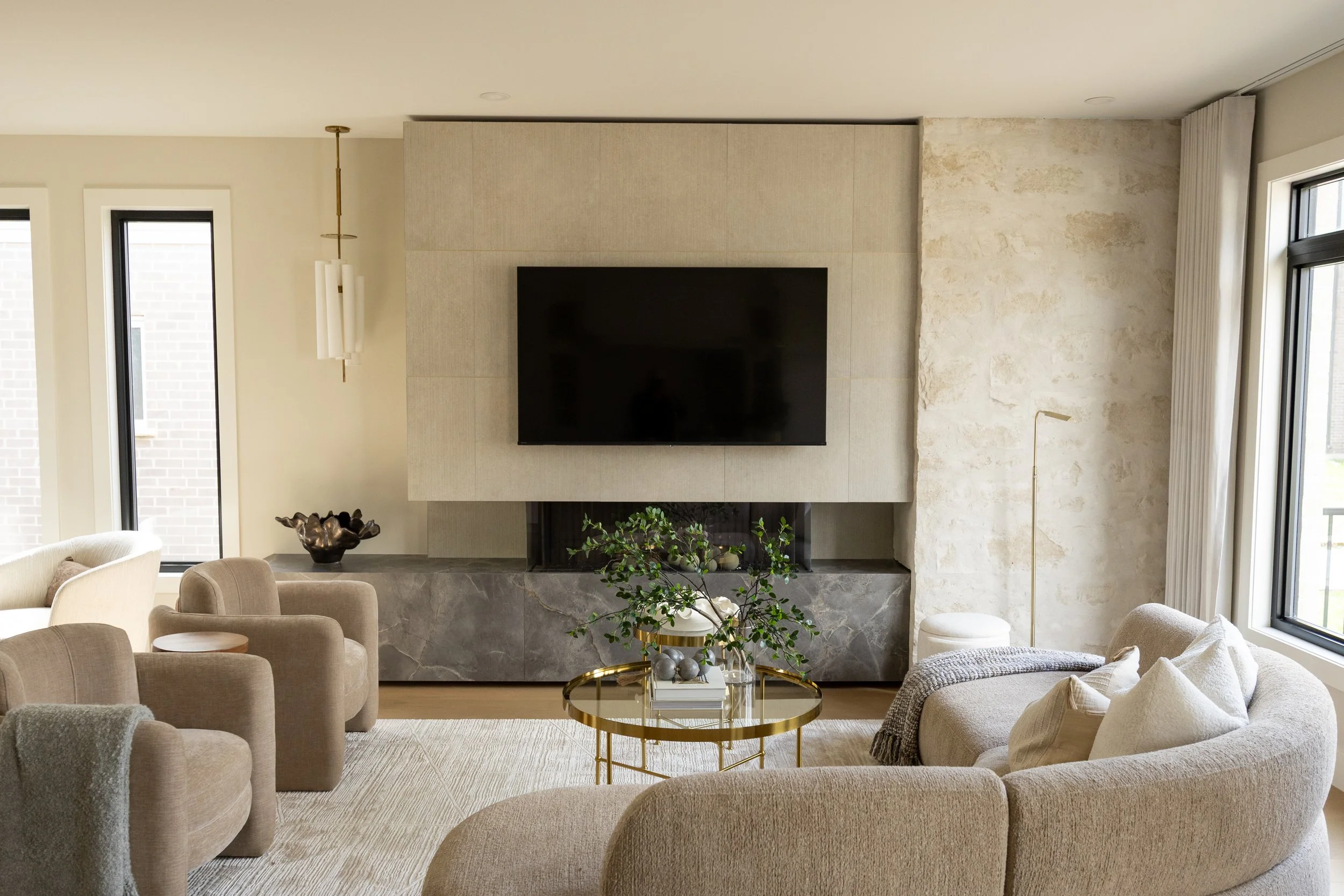 A modern living room with beige upholstered armchairs arranged around a round glass coffee table with gold accents, a fireplace with a large flat-screen TV above, and large windows letting in natural light. Decorative items include a bronze bowl and a plant.