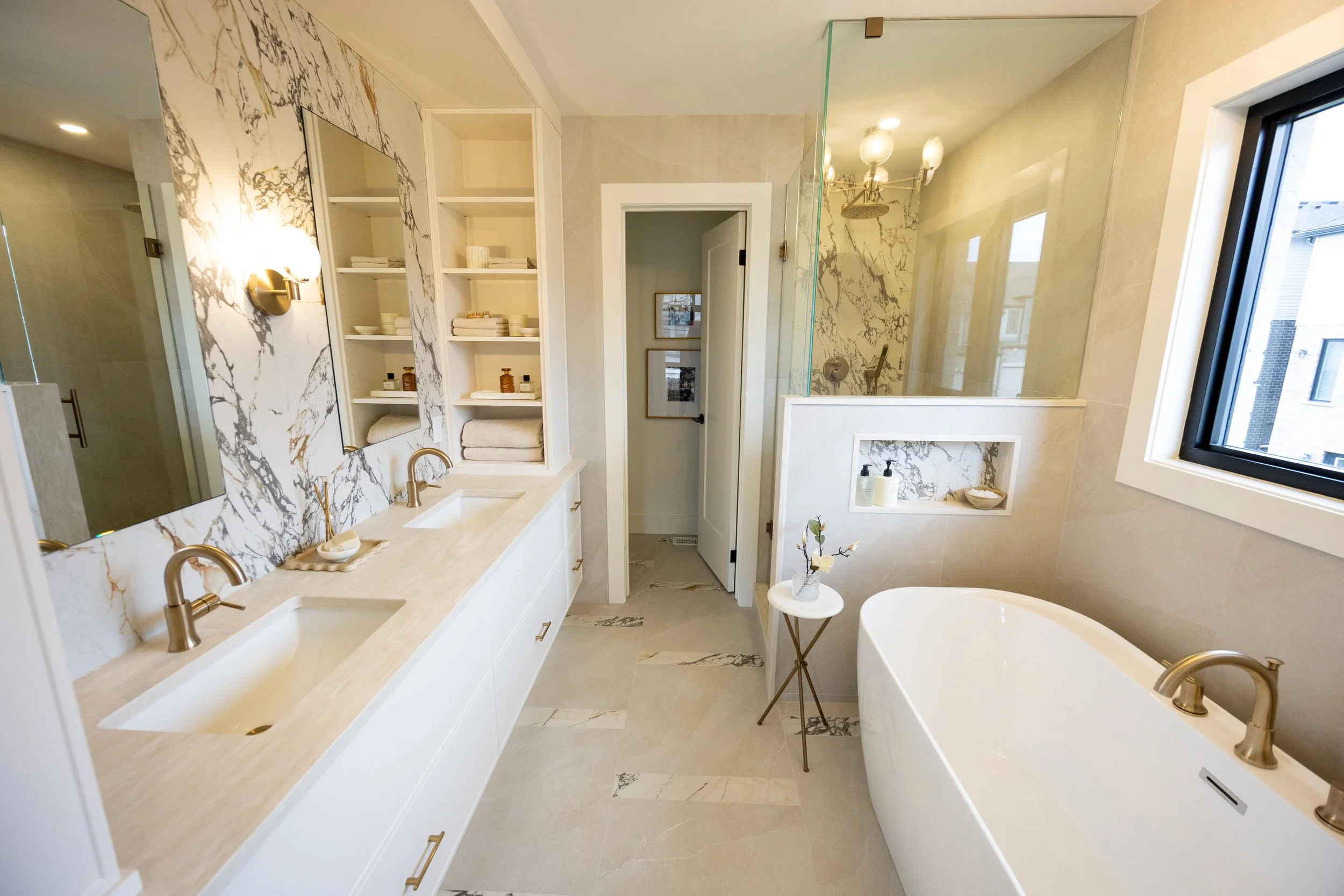Modern bathroom with a dual sink vanity, marble countertops, open shelves with towels and toiletries, a freestanding bathtub, and a walk-in shower with glass enclosure and marble accents. There is a window with a view of neighboring buildings.