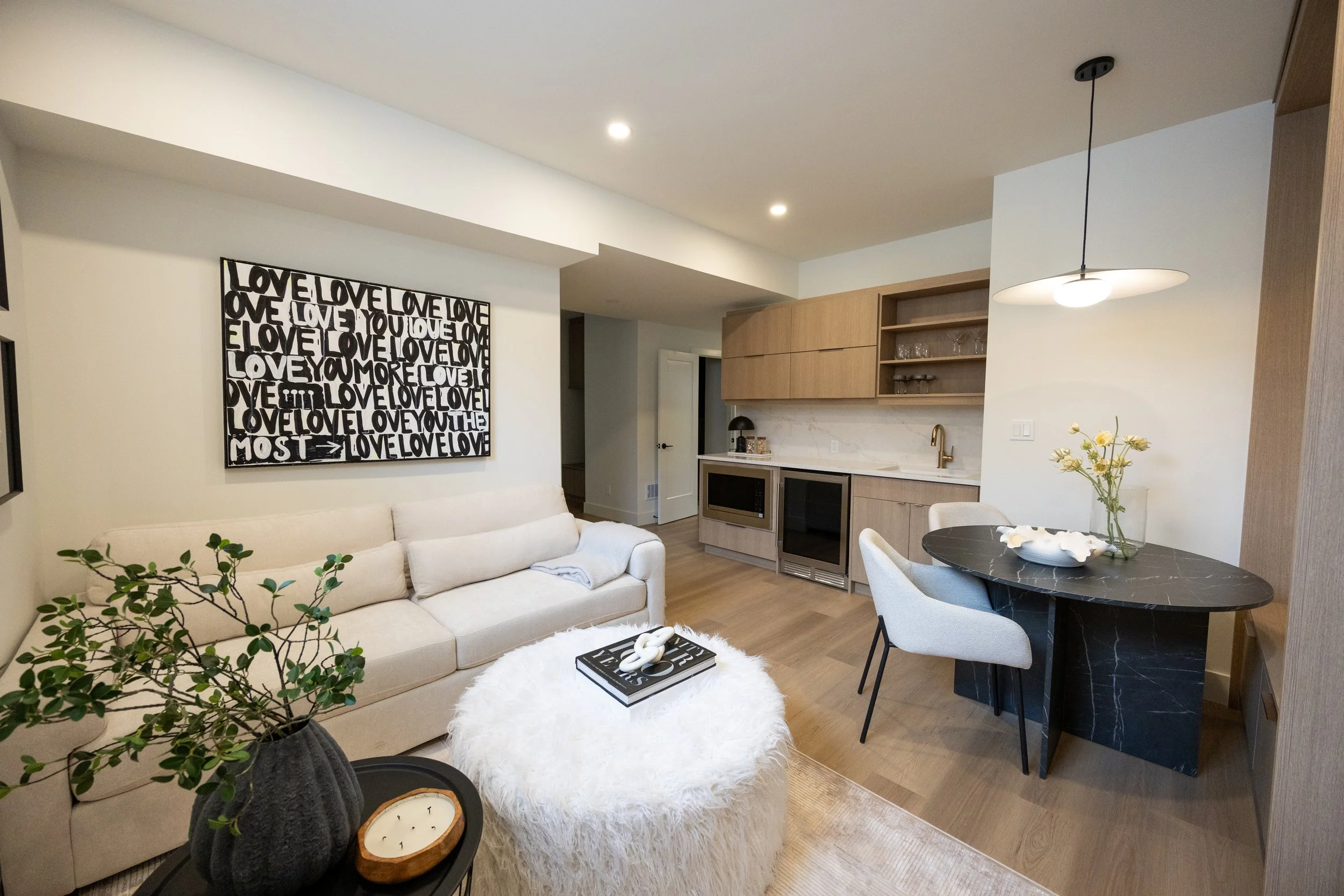 Interior view of a modern living room and kitchen area with a white sofa, black round dining table with white chairs, a black and white wall art that says "LOVE," a marble kitchen counter, wooden cabinetry, a hanging light fixture, and a vase with fl
