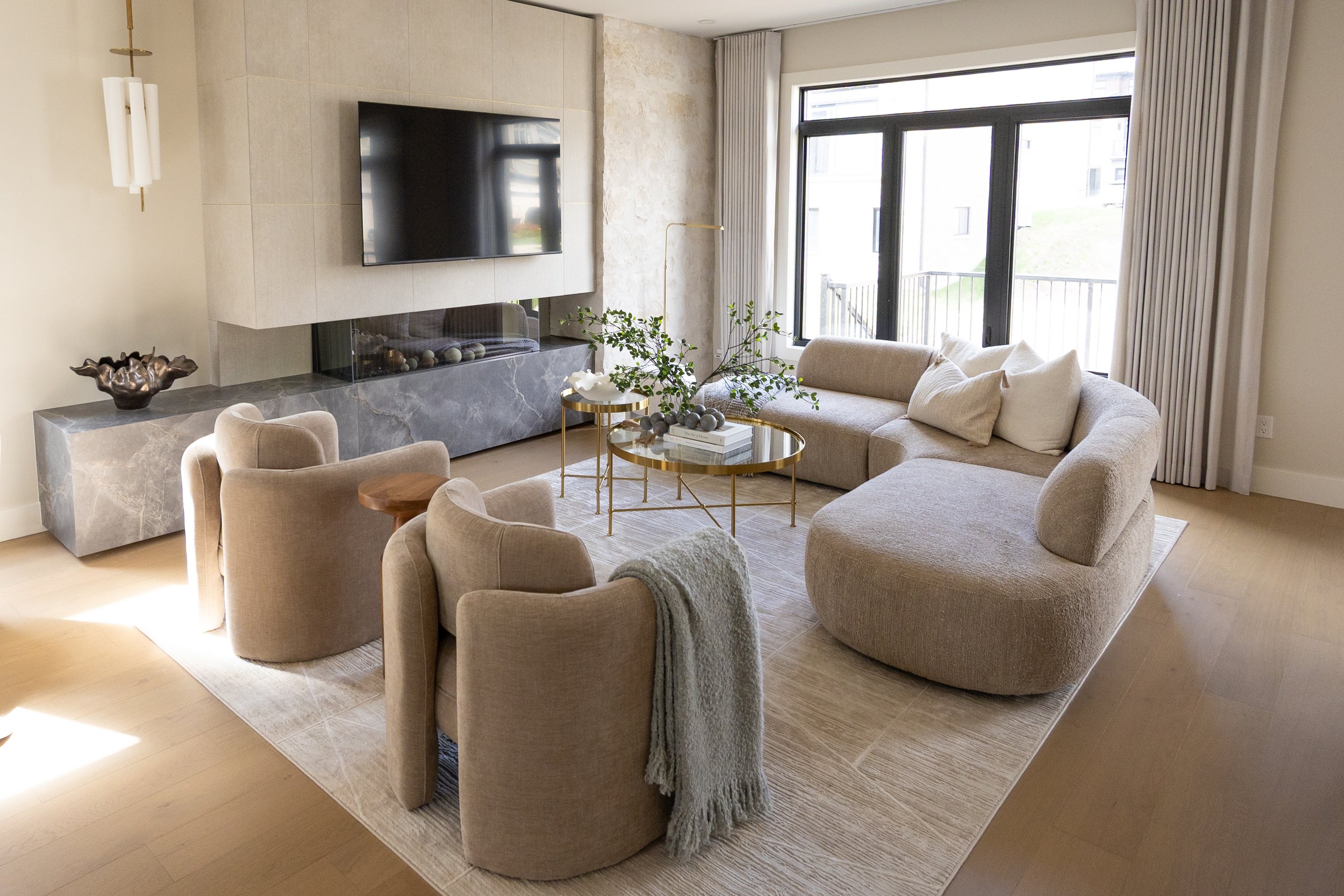 Modern living room with a beige L-shaped sofa, a glass top coffee table with decorative items, a wall-mounted flat-screen TV, large windows with white curtains, a stone fireplace, and a beige armchair with a throw blanket.
