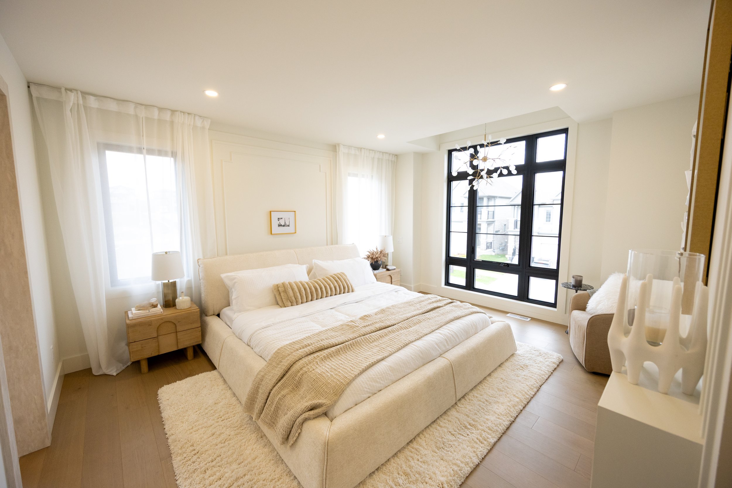 A bright, airy bedroom with a large beige upholstered bed, white bedding, and decorative pillows. There are two wooden nightstands with lamps, a framed black-and-white picture on the wall, and a large window with sheer white curtains. The room featur